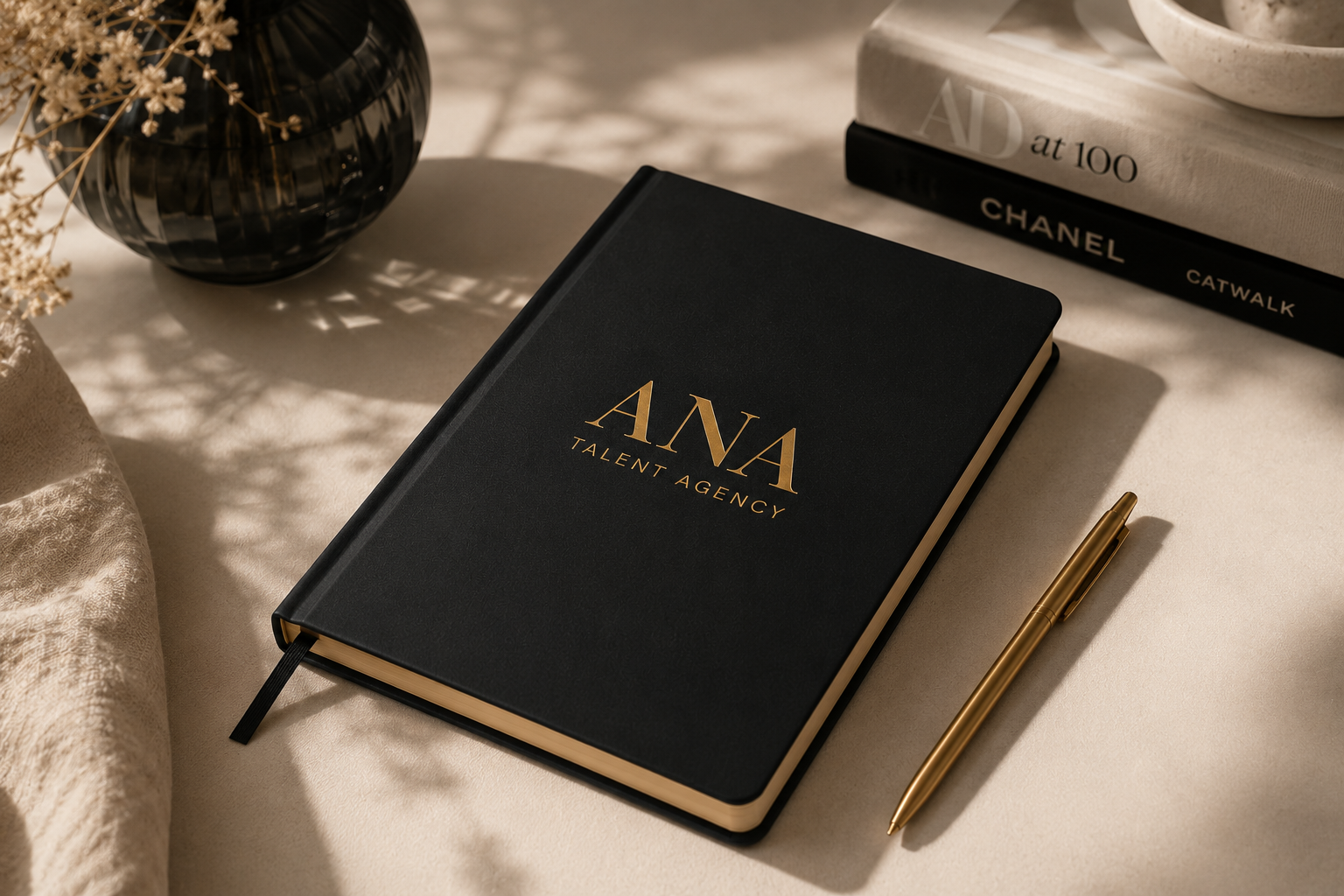 A black notebook with gold lettering reading "ANA Talent Agency" on a desk, accompanied by a gold pen, a black vase with dried flowers, and books including "Art 100" and "Chanel Catwalk."