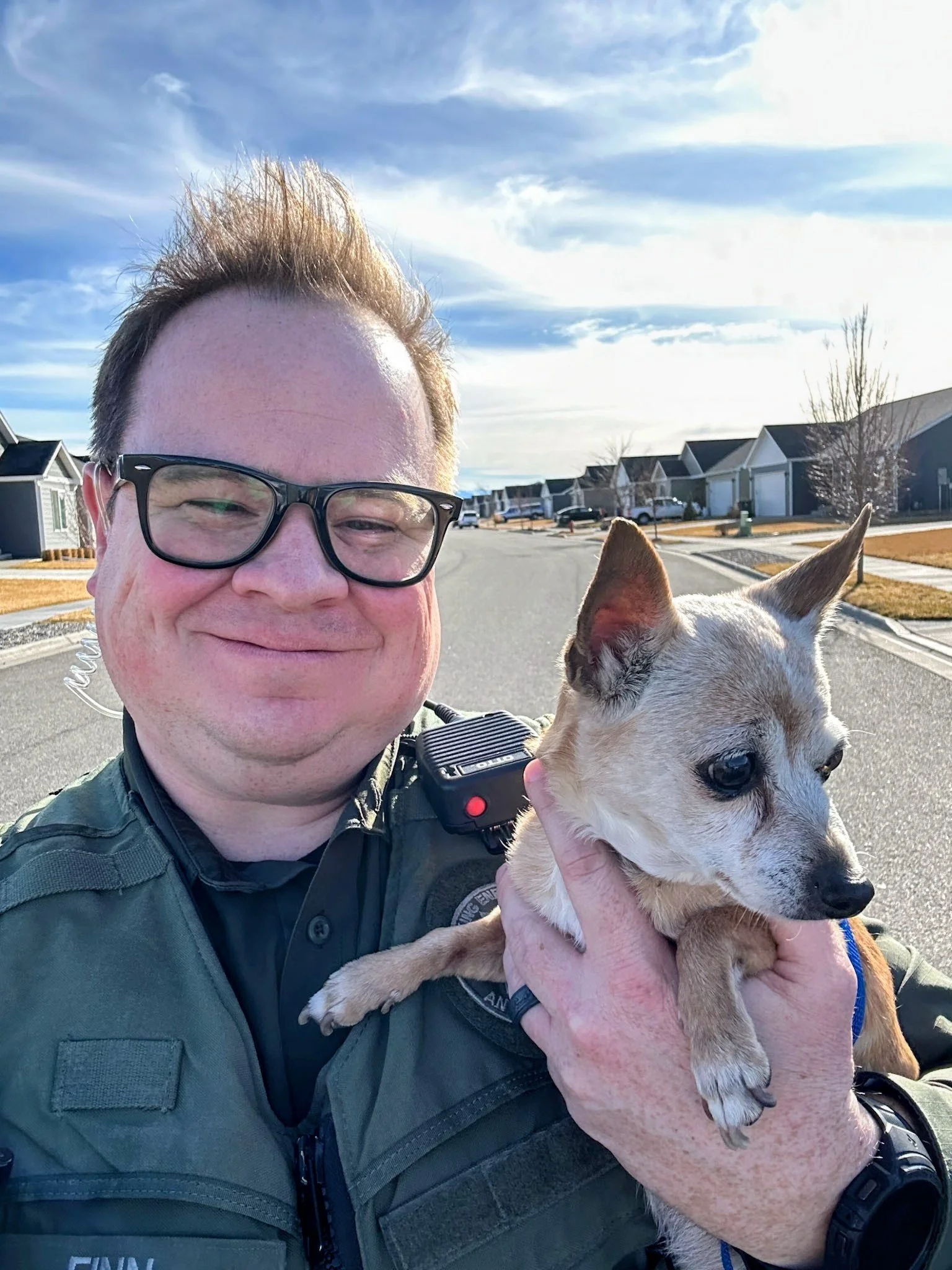 A man with glasses smiling while holding a small, light-colored dog on a sunny suburban street with houses and trees in the background.