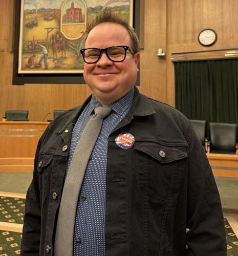 A man with glasses, smiling, wearing a black jacket, gray tie, and blue shirt, standing in a room with wooden paneling and a clock on the wall.