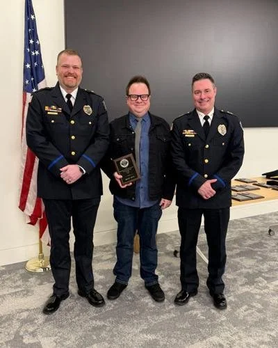 Three men standing indoors; two are police officers in uniform, and one is holding an award plaque, with an American flag in the background.