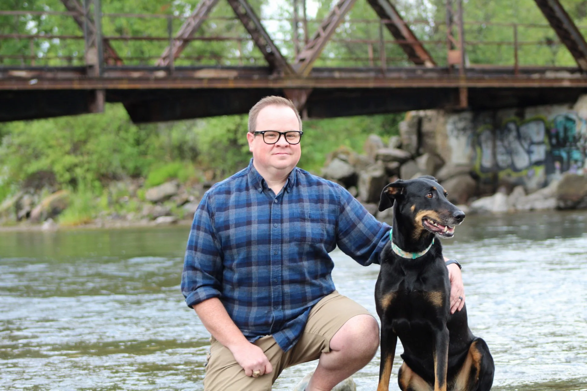 A man with glasses in a plaid shirt kneeling by a black and tan dog in a river with a bridge and green trees in the background.