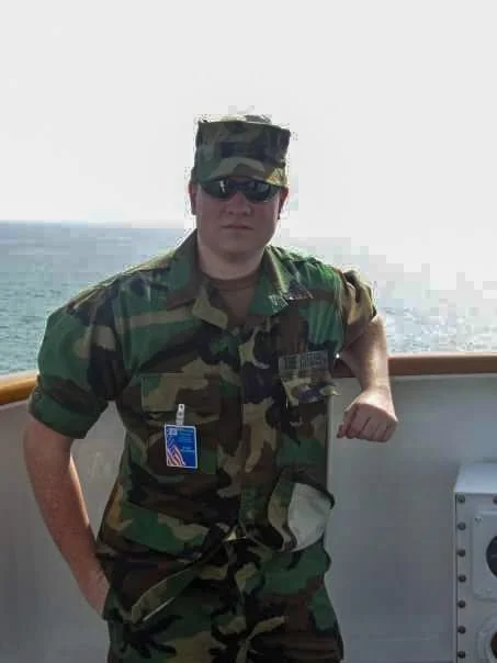 Person wearing camouflage military uniform and sunglasses standing on a boat near the ocean