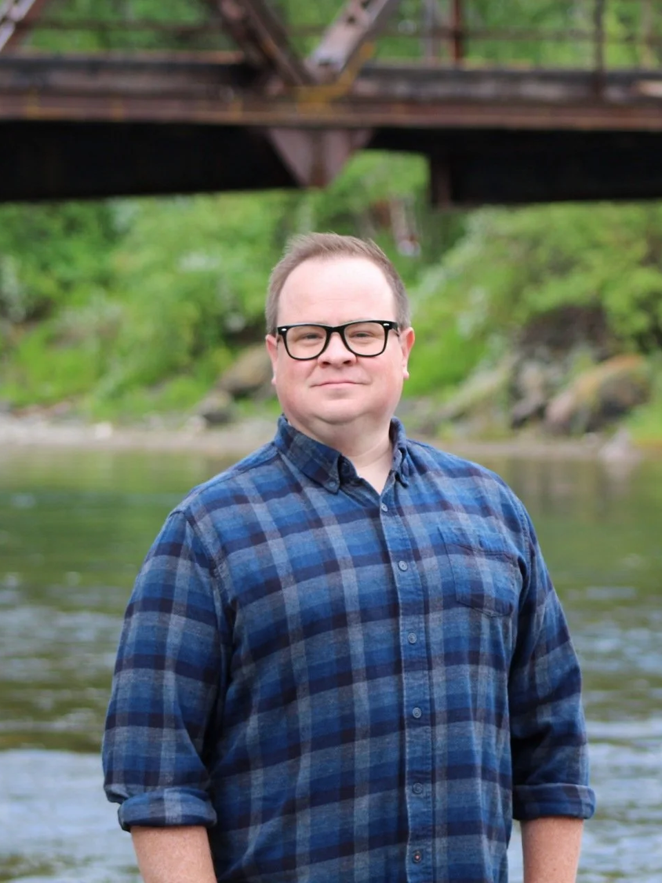 Man wearing glasses and a blue plaid shirt standing in front of a river with greenery and a bridge overhead.