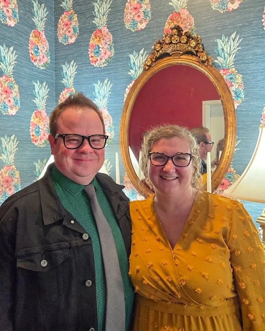 A smiling man and woman wearing glasses stand together in front of a floral pineapple-patterned wallpaper and an oval mirror with ornate gold frame. The woman is wearing a yellow dress, and the man is wearing a green shirt, gray tie, and black jacket.