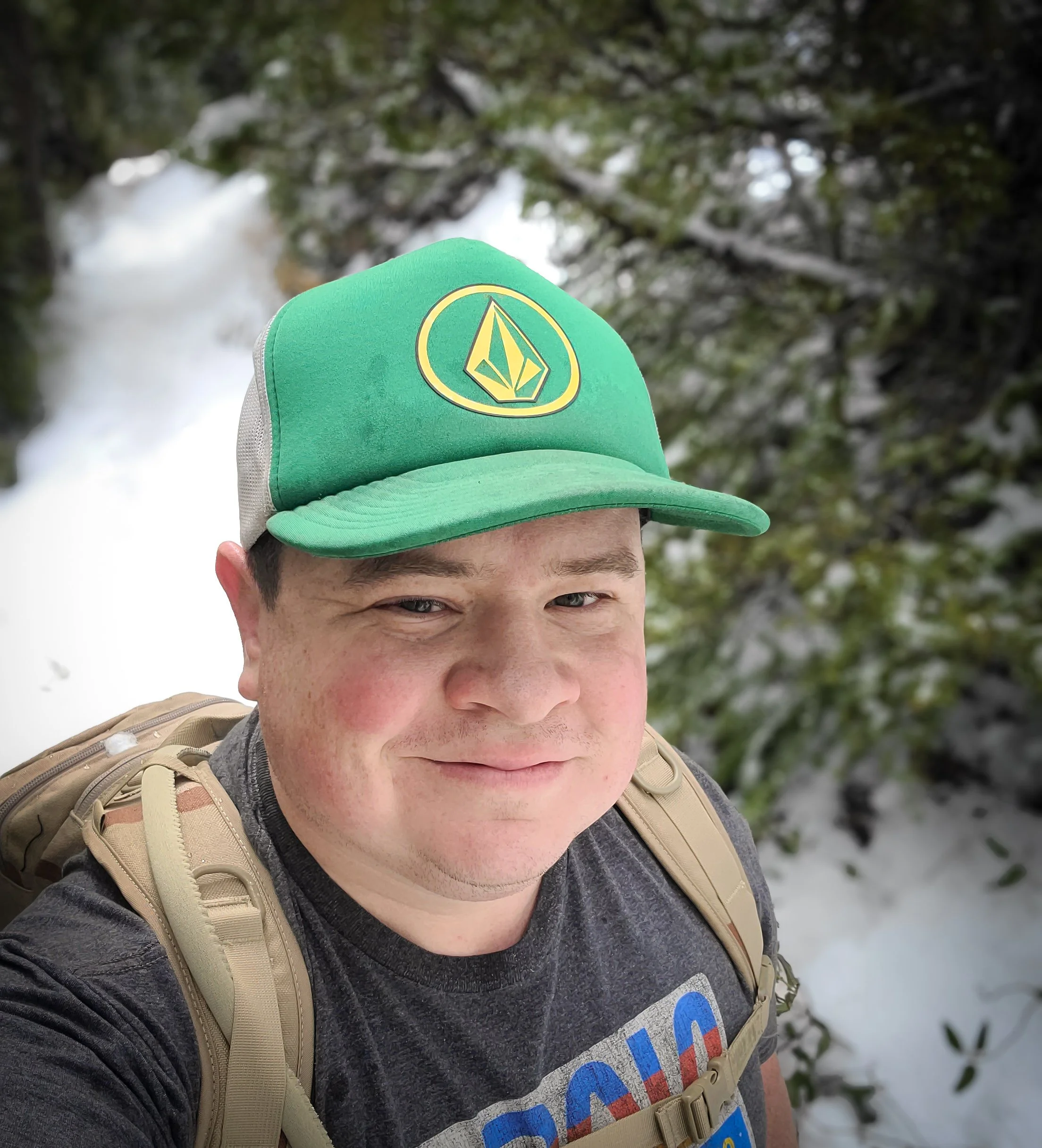 A man taking a selfie outdoors during winter with snow-covered trees in the background. He is wearing a black t-shirt, a beige backpack, and a green cap with a yellow logo. The man has a slight smile on his face.