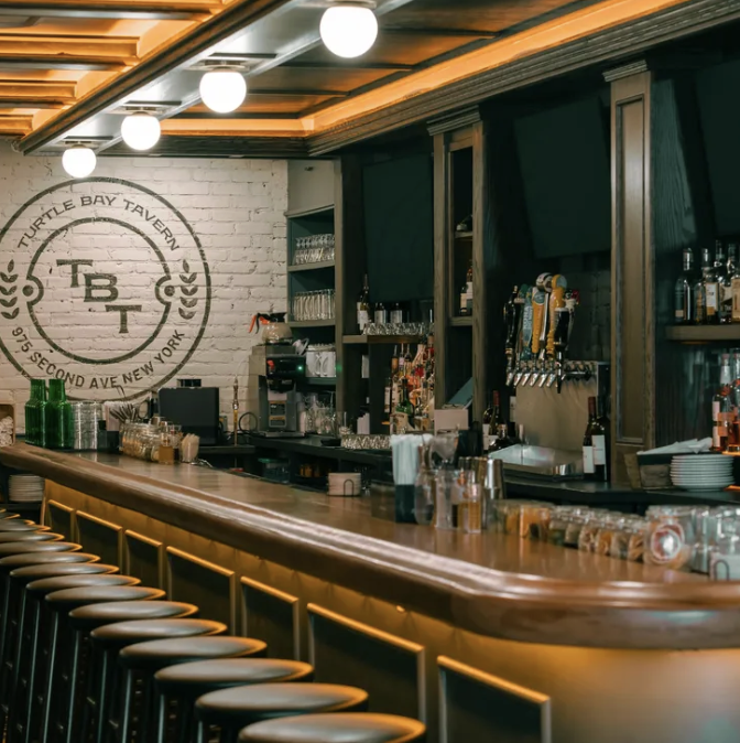 Interior of a bar at Turtle Bay Tavern in New York, showing a long wooden counter with bar stools, shelves with glasses, and beer taps.