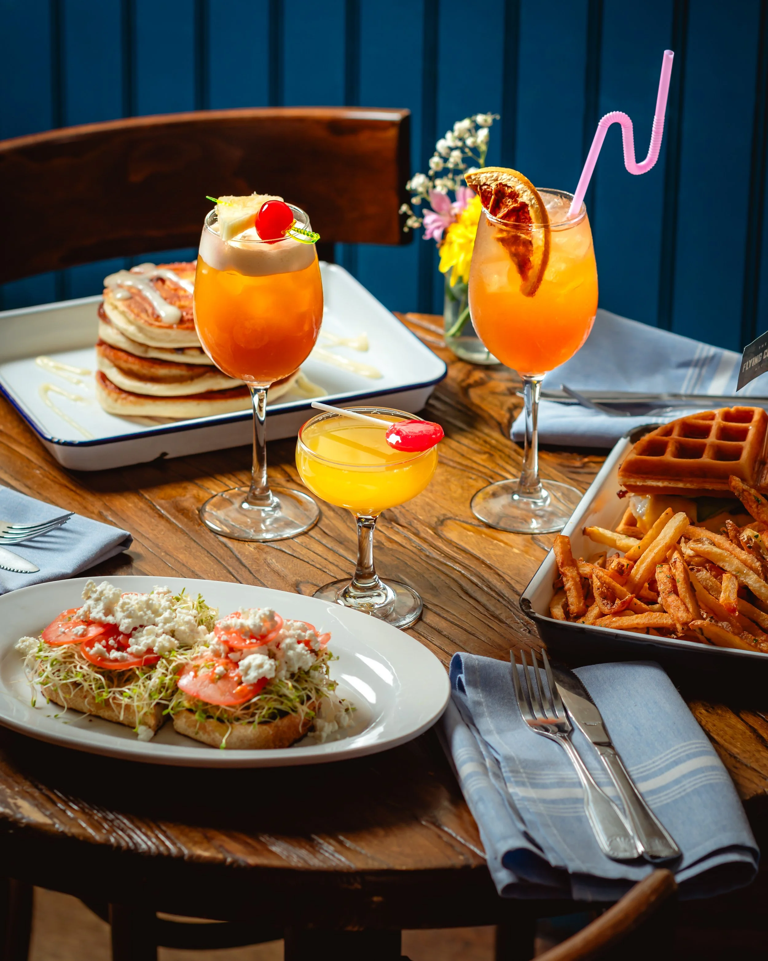 Table with brunch food and drinks, including a plate of chicken and waffles, a salad, and two colorful cocktails with fruit garnishes and pink straws, set with a fork and knife on a blue napkin.