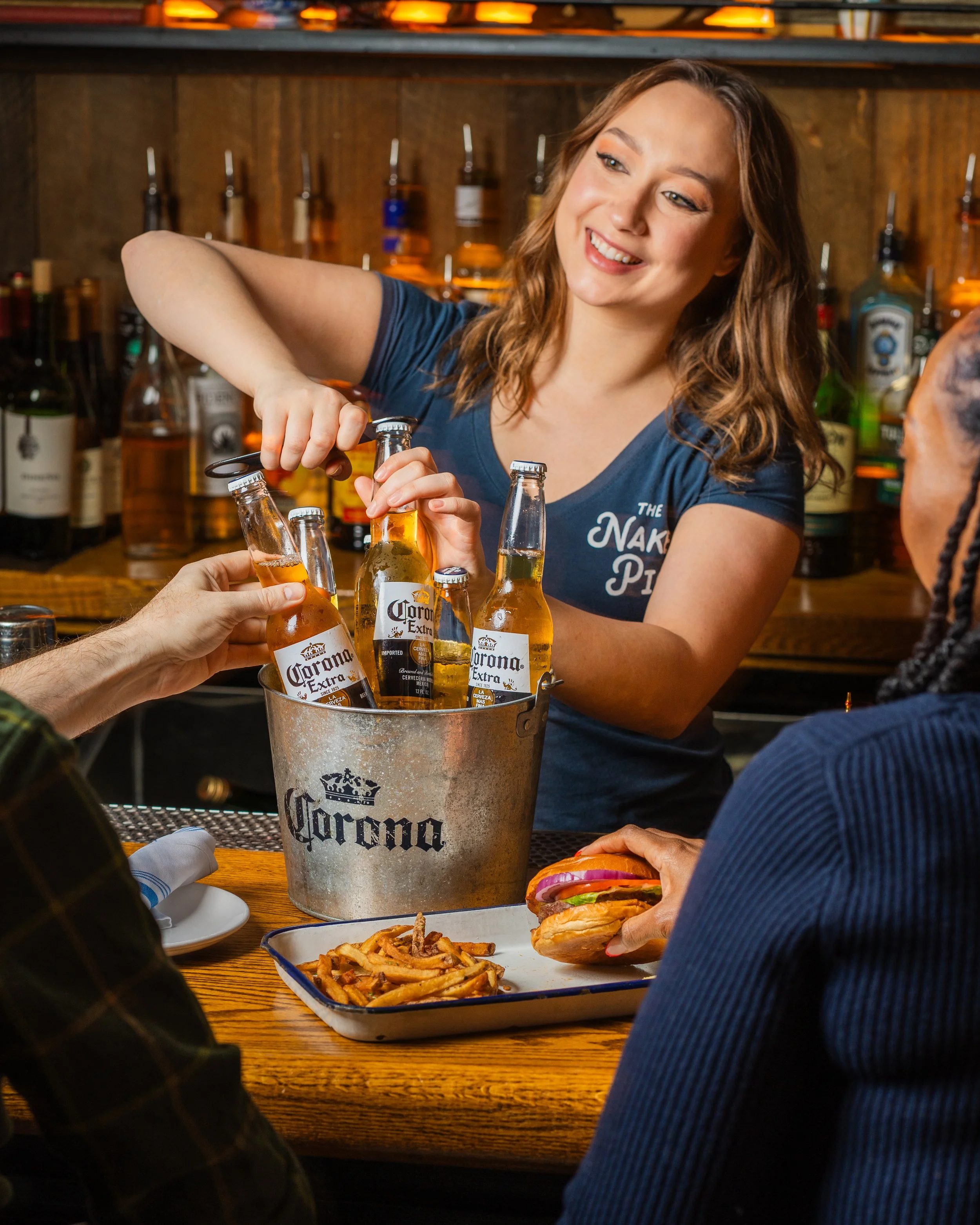 A woman with brown wavy hair smiling and preparing to serve drinks at a bar. She is wearing a blue t-shirt, and there are bottles of alcohol in the background. On the table, there are plates of food including fries, a burger with lettuce, tomato, and onion, and a bucket of Corona beer bottles.