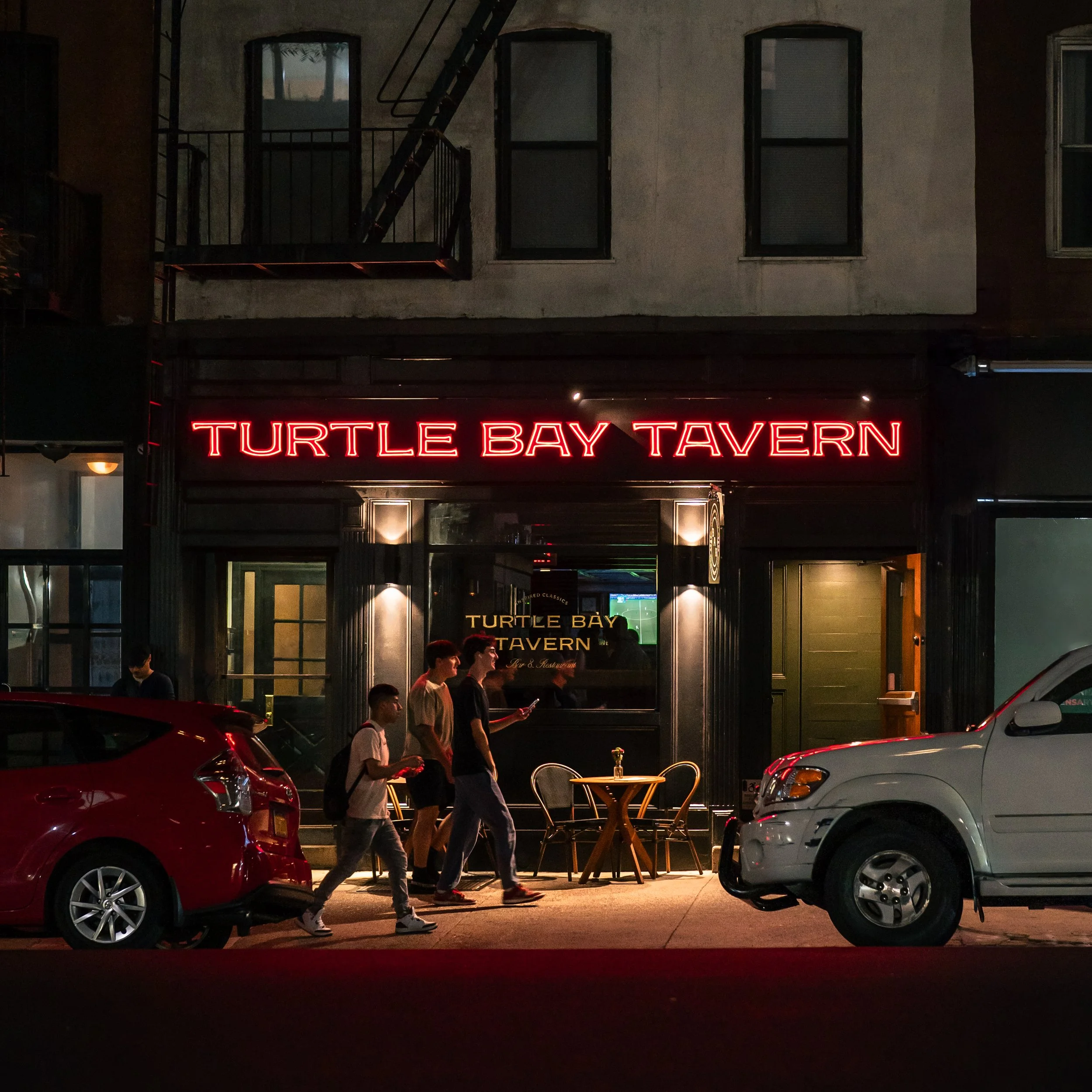 Nighttime scene of a street view outside Turtle Bay Tavern, with people walking on the sidewalk, cars parked on the street, and neon signage displaying the tavern's name.