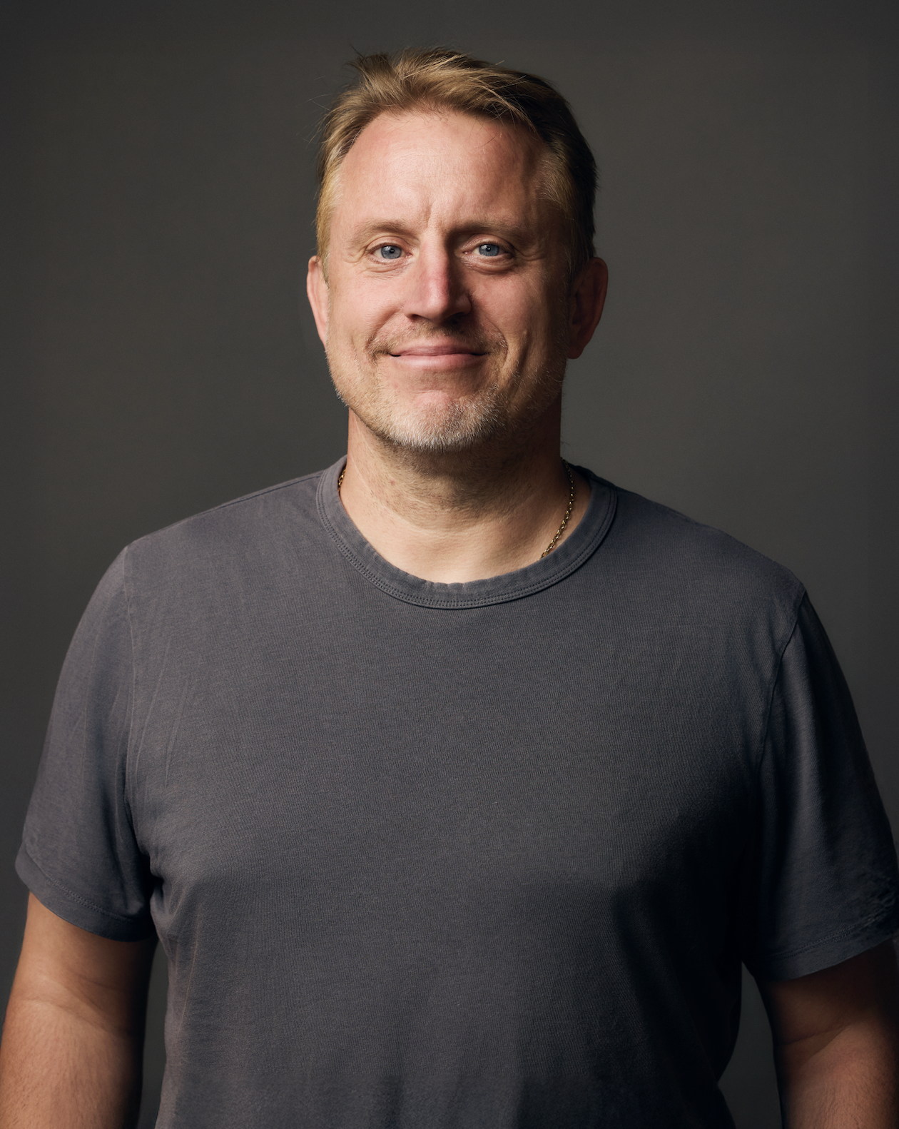 A middle-aged man with blond hair, light facial hair, blue eyes, and a slight smile, wearing a dark gray t-shirt and a gold chain, standing against a dark gray background.