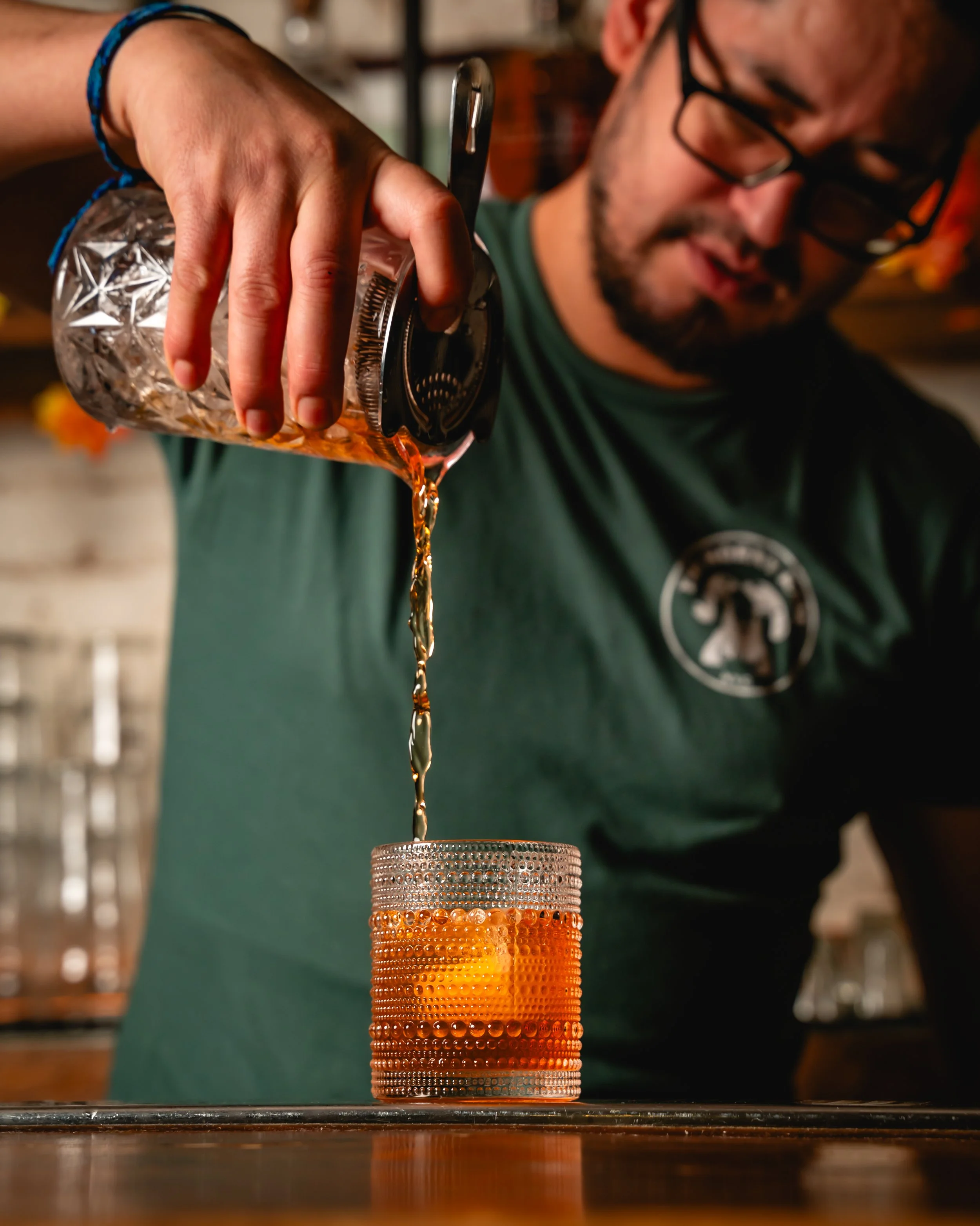 A person pouring a drink from a glass pitcher into a textured glass on a wooden surface.