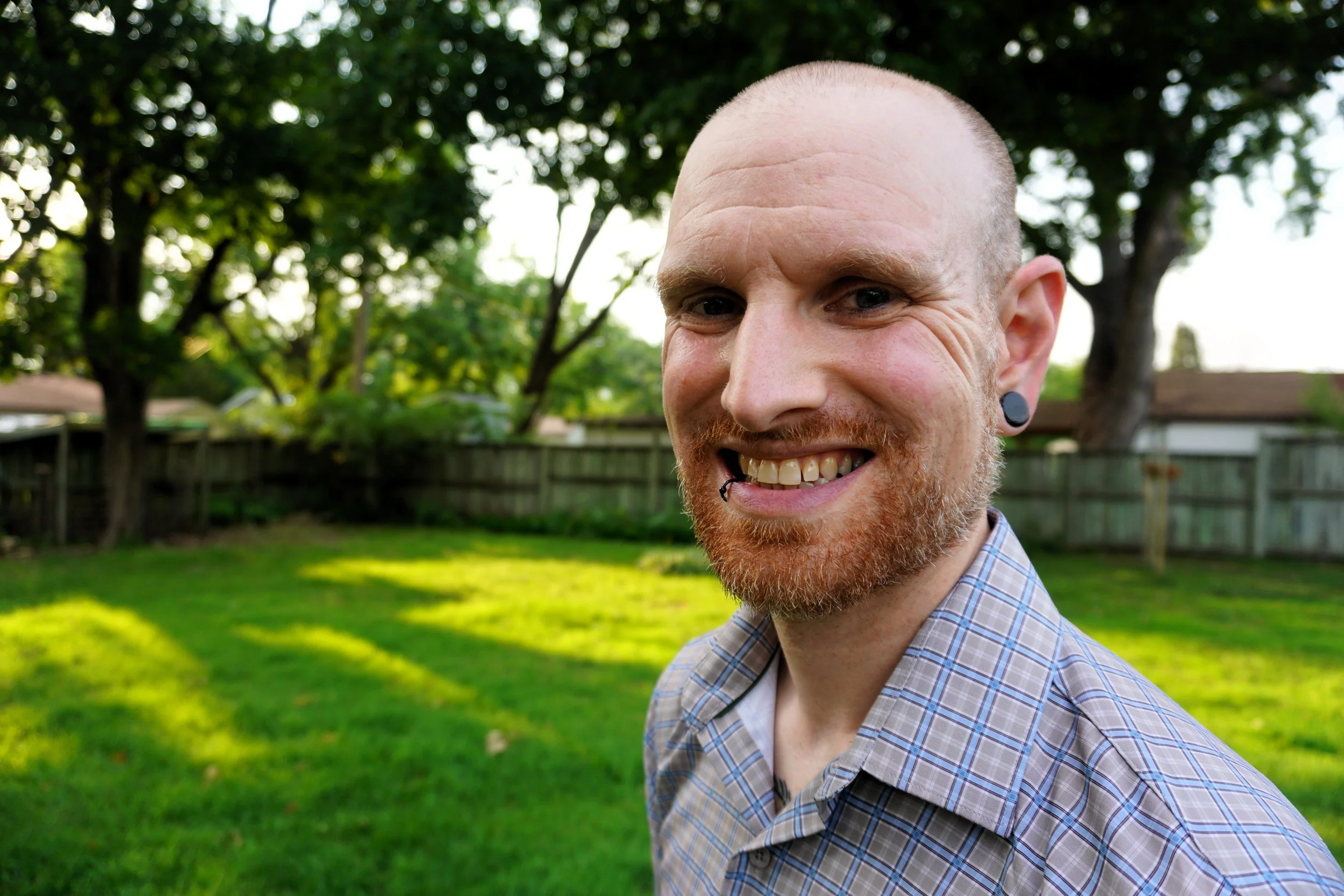 Close-up of a smiling man with a bald head, red beard, black earrings, wearing a plaid shirt, outdoors with green trees and a wooden fence in the background.