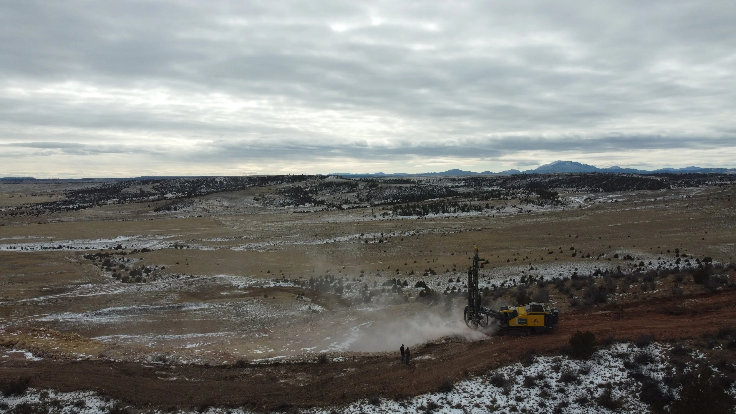 A landscape of rolling hills with patchy snow, under a cloudy sky. There is a large yellow drilling machine in the foreground, with a few people nearby, suggesting construction or drilling activity.