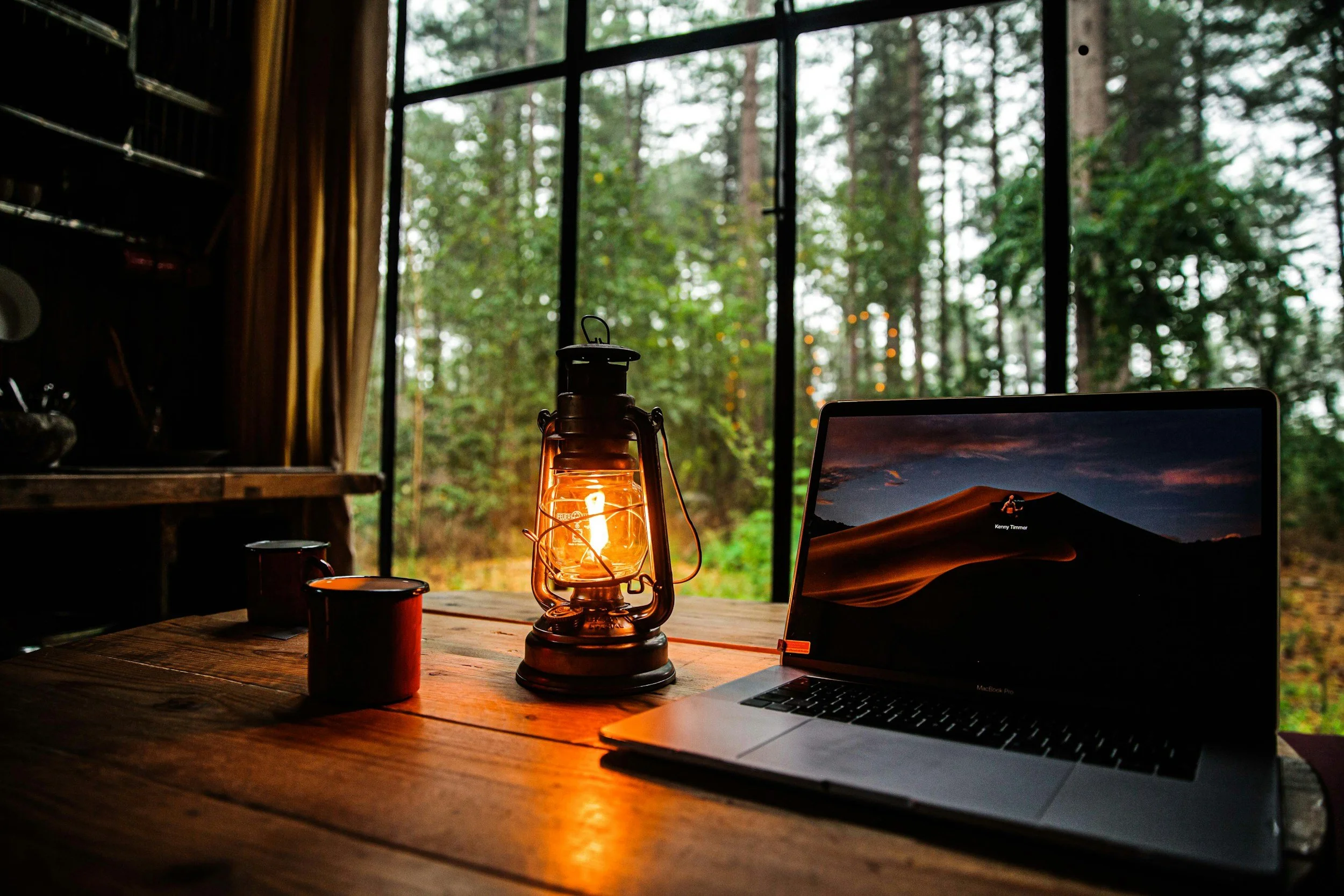 Wooden table with a vintage lantern lit, a laptop, and a red mug inside a cozy cabin with a large window showing a forest outside.