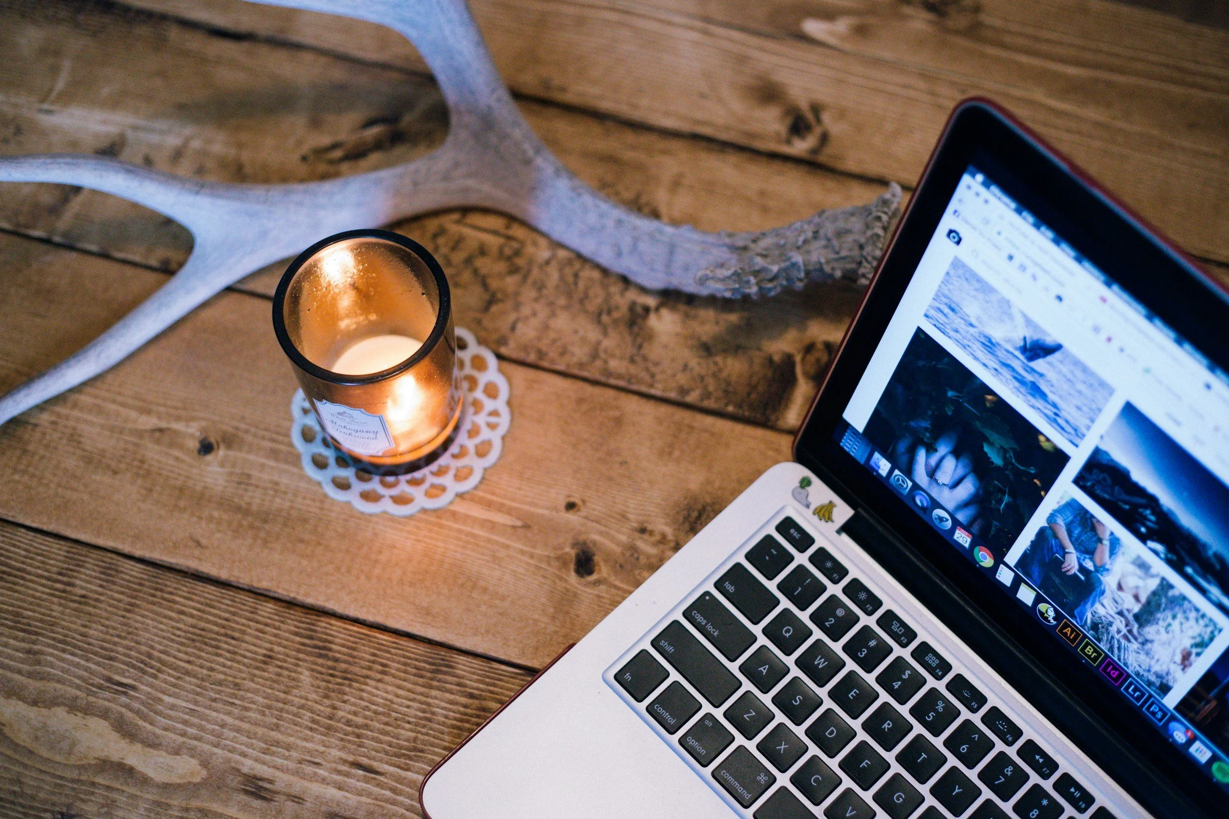 A laptop on a wooden table displaying a photo website, with a lit candle in a glass holder and a decorative antler in the background.