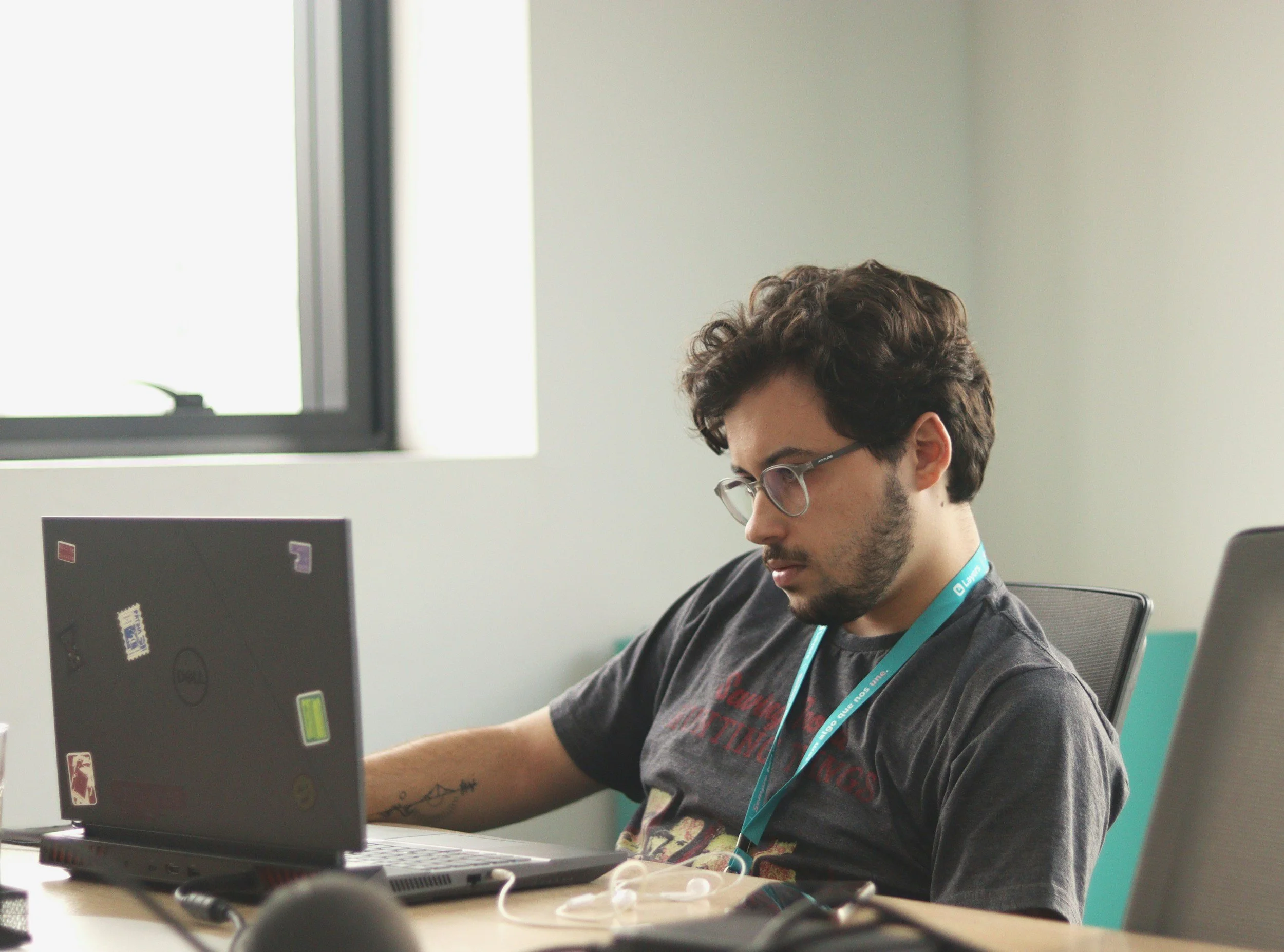 A young man with glasses and curly hair sitting at a desk working on a laptop in an office.