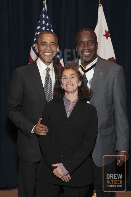 Three smiling people, two men and one woman, stand together in front of American and California flags, with dark curtains behind them.