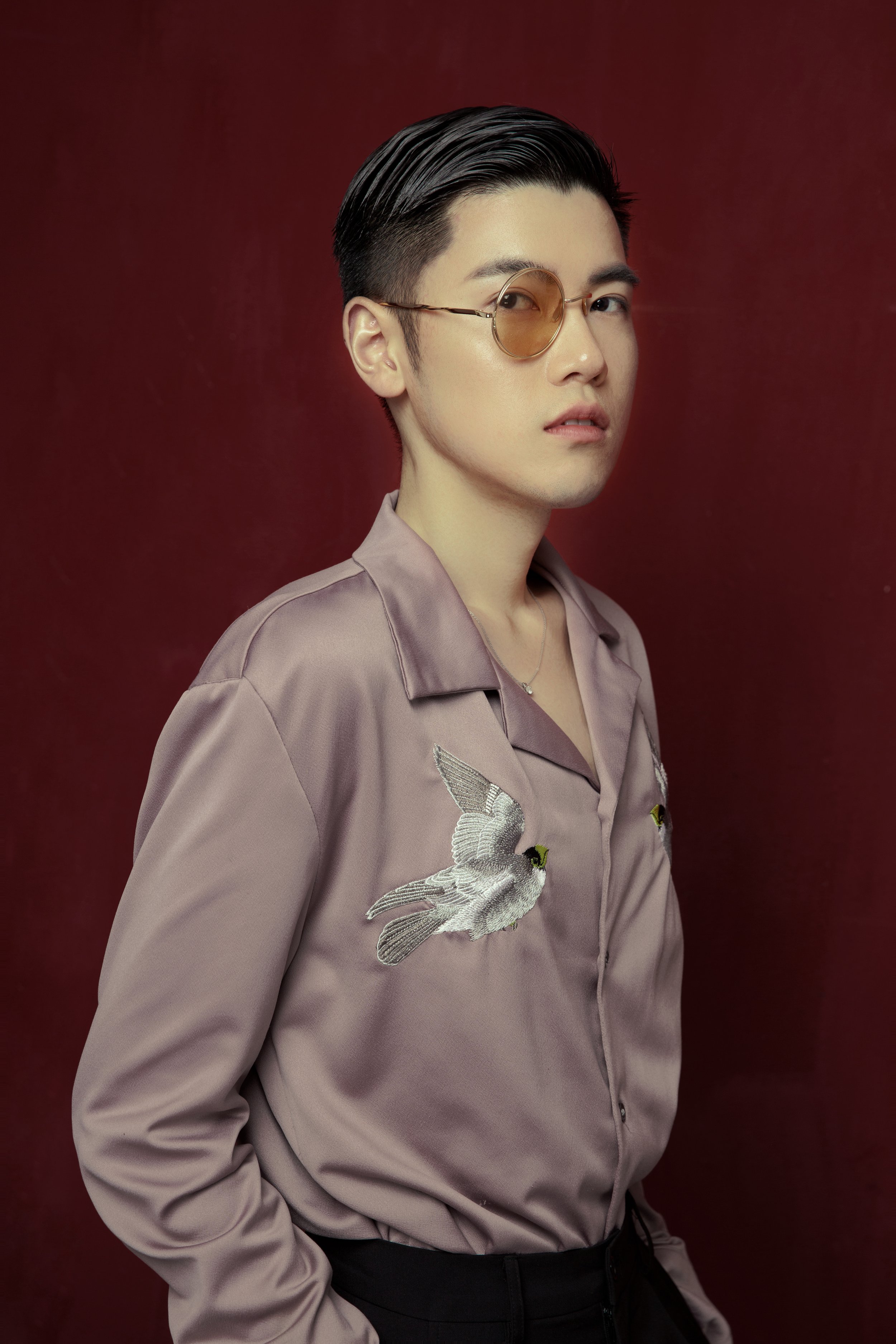 A person with short dark hair, wearing round tinted glasses and a satin shirt with embroidered birds, standing against a red background.
