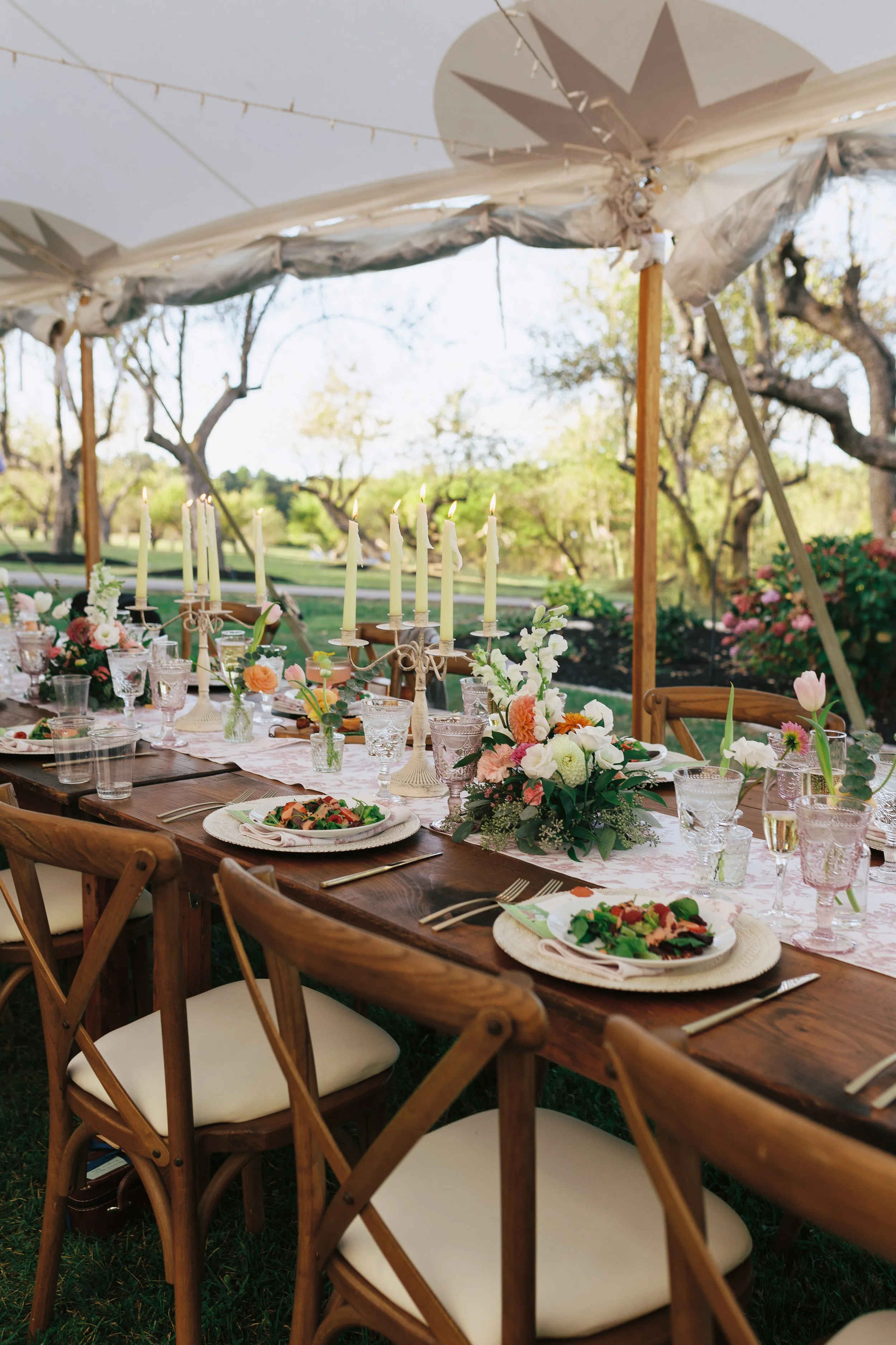 A long wooden outdoor dining table set for an event, decorated with floral arrangements, candles, and glassware, under a large white canopy with string lights, in a lush garden setting.