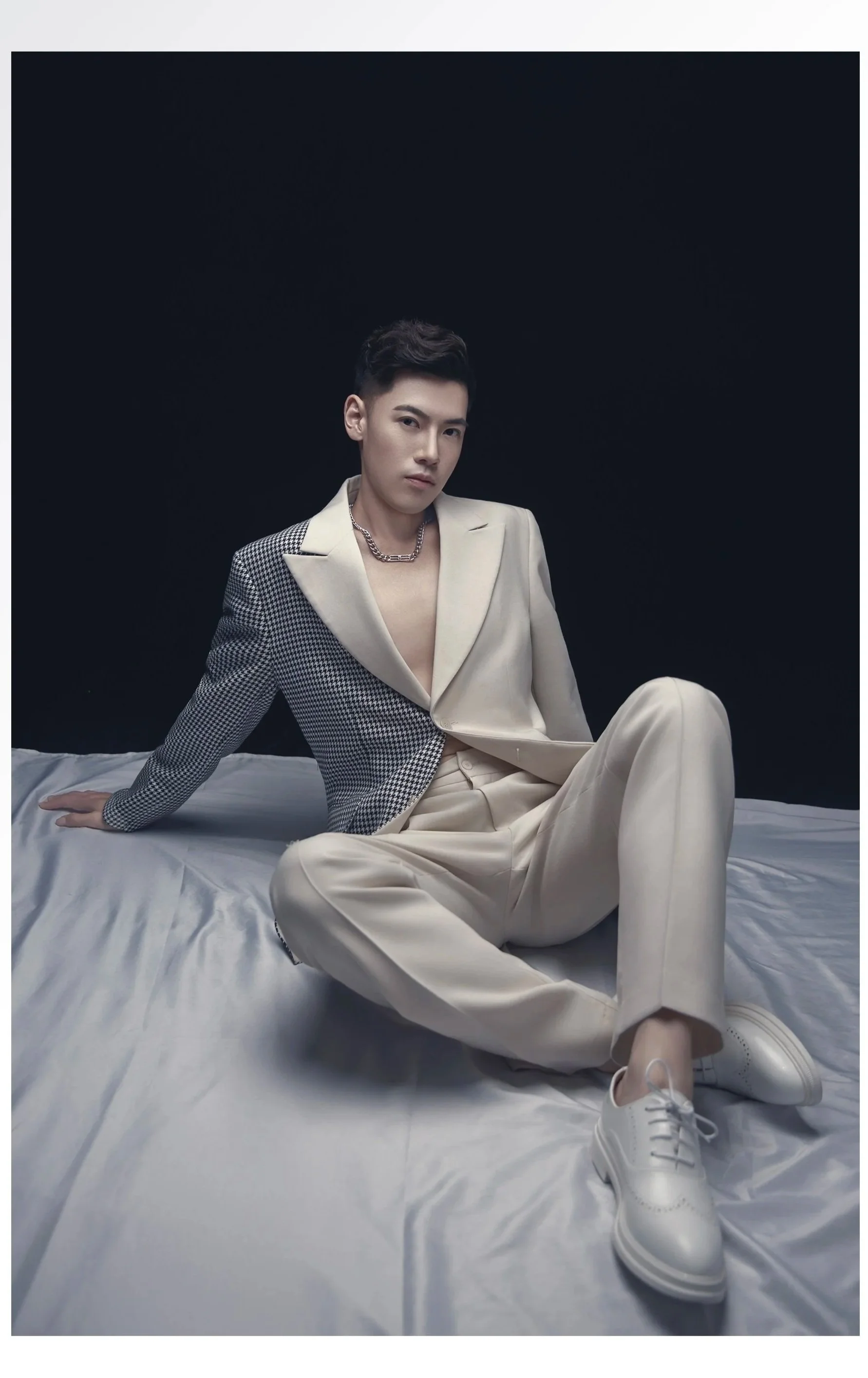 A young man with short dark hair and light skin sits on a bed against a black background. He wears a beige suit with one half featuring a checkered pattern and the other plain, an open shirt, a chain necklace, and white sneakers. He poses with one arm supporting him and the other resting on his knee, looking directly at the camera.