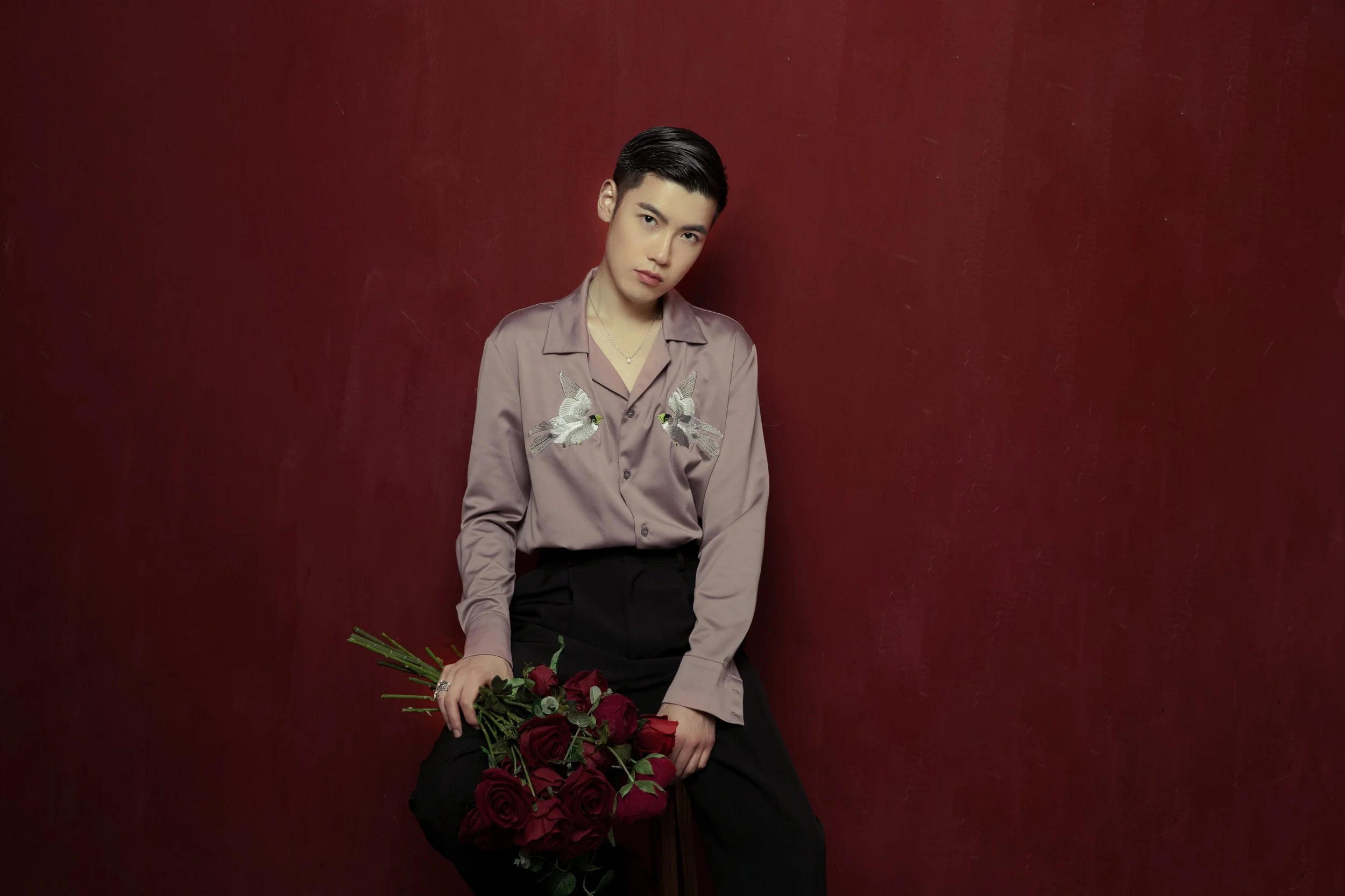 A young person with short dark hair wearing a beige satin shirt with embroidered birds, sitting against a red wall, holding a bouquet of red roses.
