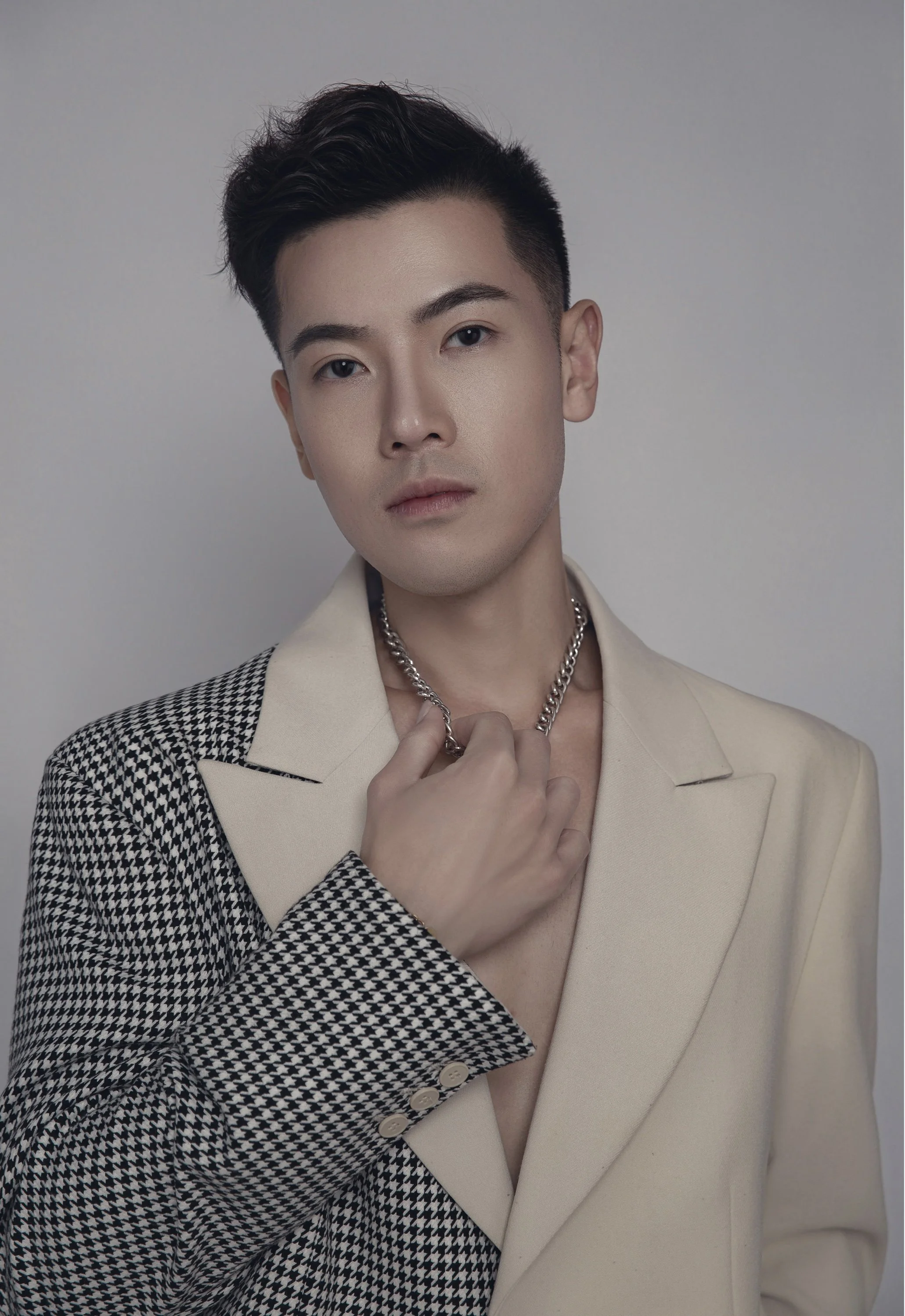A young man with dark, styled hair and light skin, wearing a light beige blazer and a houndstooth patterned shirt, holding a silver chain necklace, in front of a plain gray background.