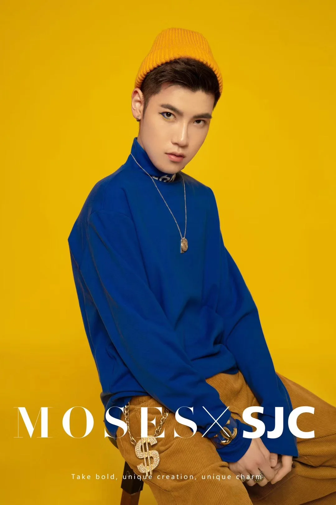 A young man is sitting against a yellow background, wearing a yellow beanie, blue shirt, brown pants with a dollar sign chain, and a necklace, with a confident expression.