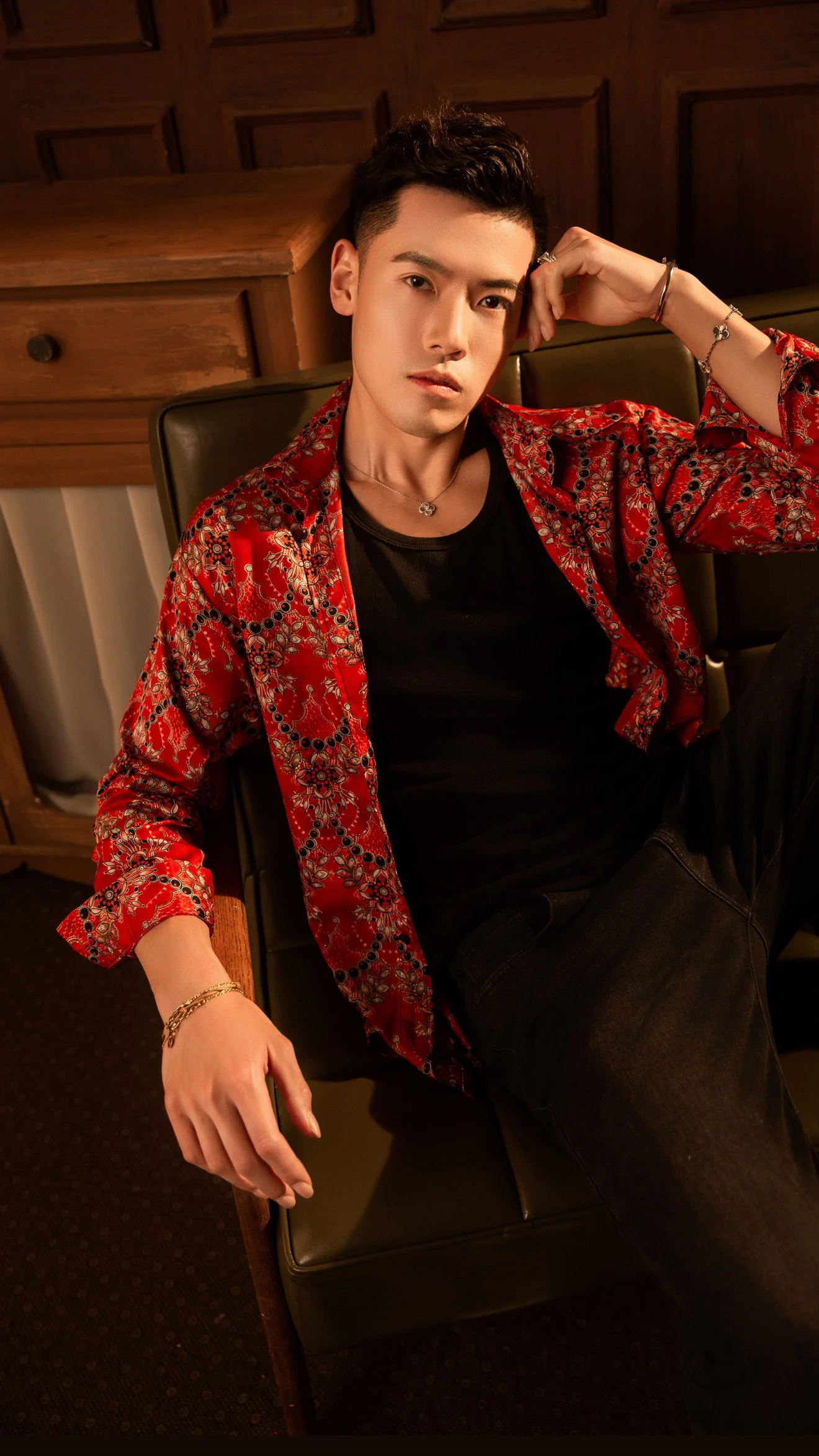 Young man with dark hair, wearing a red patterned shirt over a black T-shirt, sitting in a black leather chair in a dimly lit room with wooden paneling on the wall.