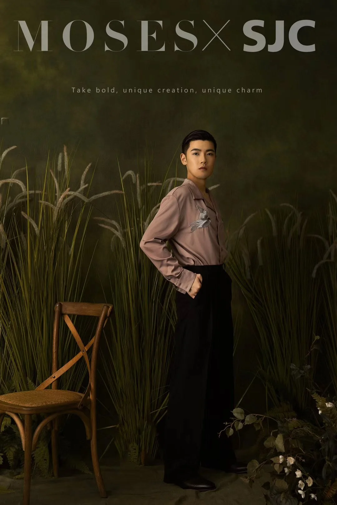 A person standing in front of a dark background with tall grass and plants, wearing a brown silky shirt with embroidery and black pants, with a wooden chair nearby.