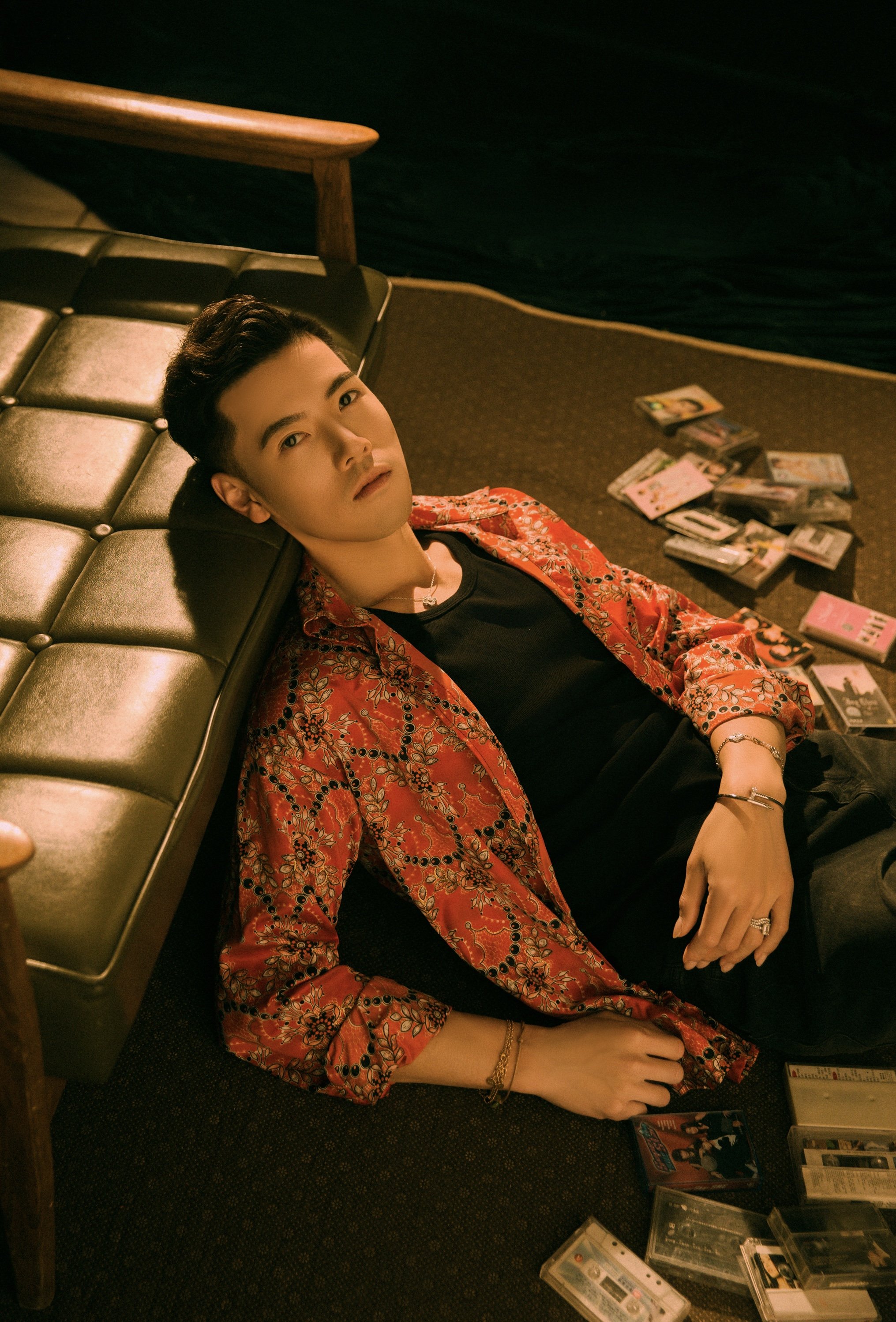 A young man with black hair lying on a dark carpeted floor, wearing a red patterned shirt over a black t-shirt, surrounded by cassette tapes and cassette cases. He is resting his head on a leather bench, looking at the camera.
