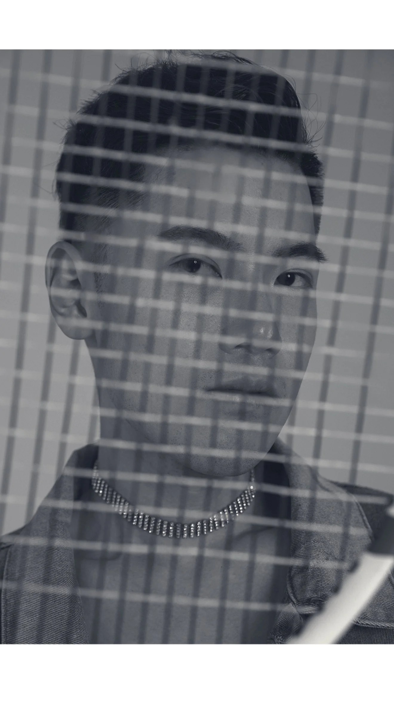 A black-and-white photo of a young man with short hair, wearing a chain necklace and a collared shirt, seen through a grated surface with horizontal and vertical lines.