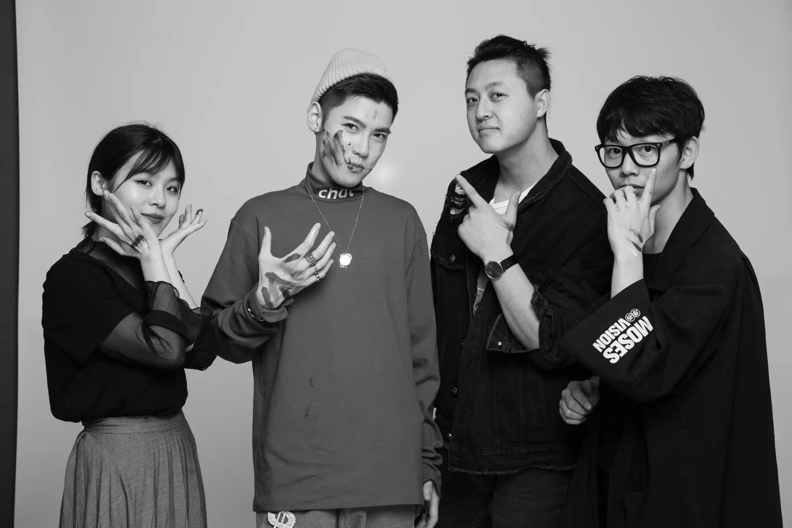 Four young adults, two men and two women, posing together in a black and white photo against a plain background. They are making playful hand gestures and have paint or makeup on their faces and hands.