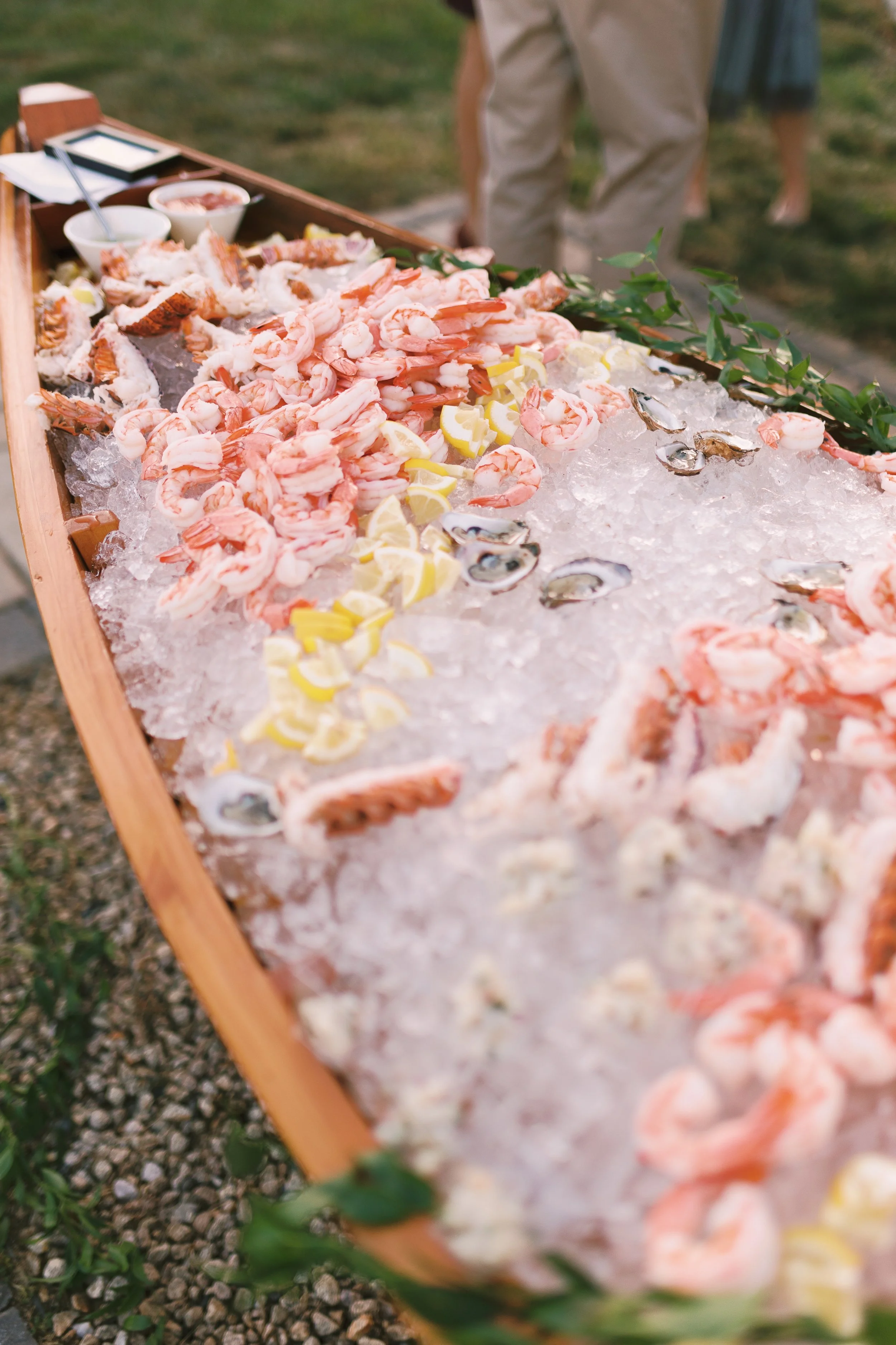A wooden seafood display table covered with crushed ice, featuring shrimp, oysters, shellfish, lemon slices, and small bowls of dipping sauces, with greenery decoration along the edge.
