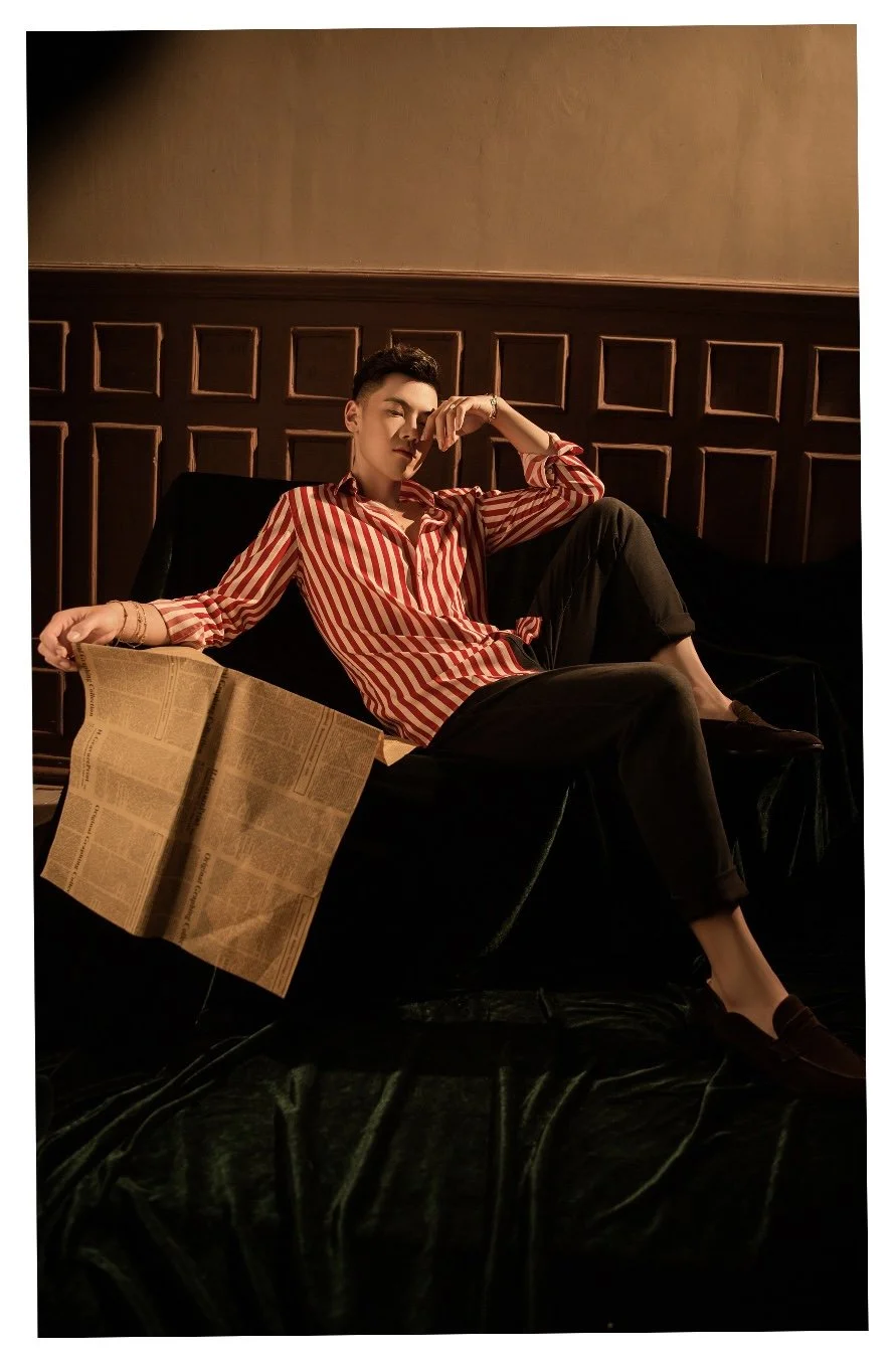 A young man with styled hair, wearing a red and white striped shirt and black pants, is sitting on a black sofa in a dimly lit room. He has one arm resting on his forehead and is holding a newspaper in his other hand, with a relaxed and contemplative