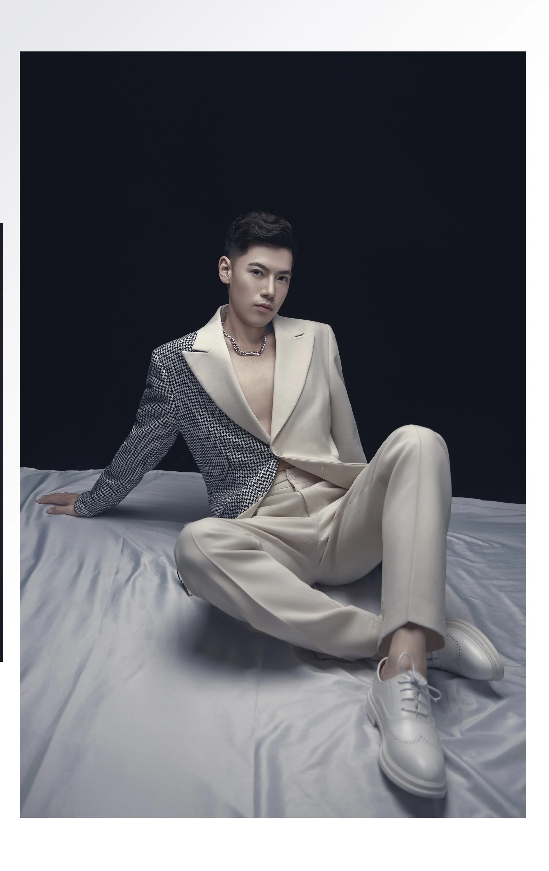 A young man with short dark hair sitting on a bed against a black background. He is wearing a cream-colored suit jacket with a checkered pattern on the right side, cream pants, and white sneakers. He has a silver chain necklace and a serious expressi