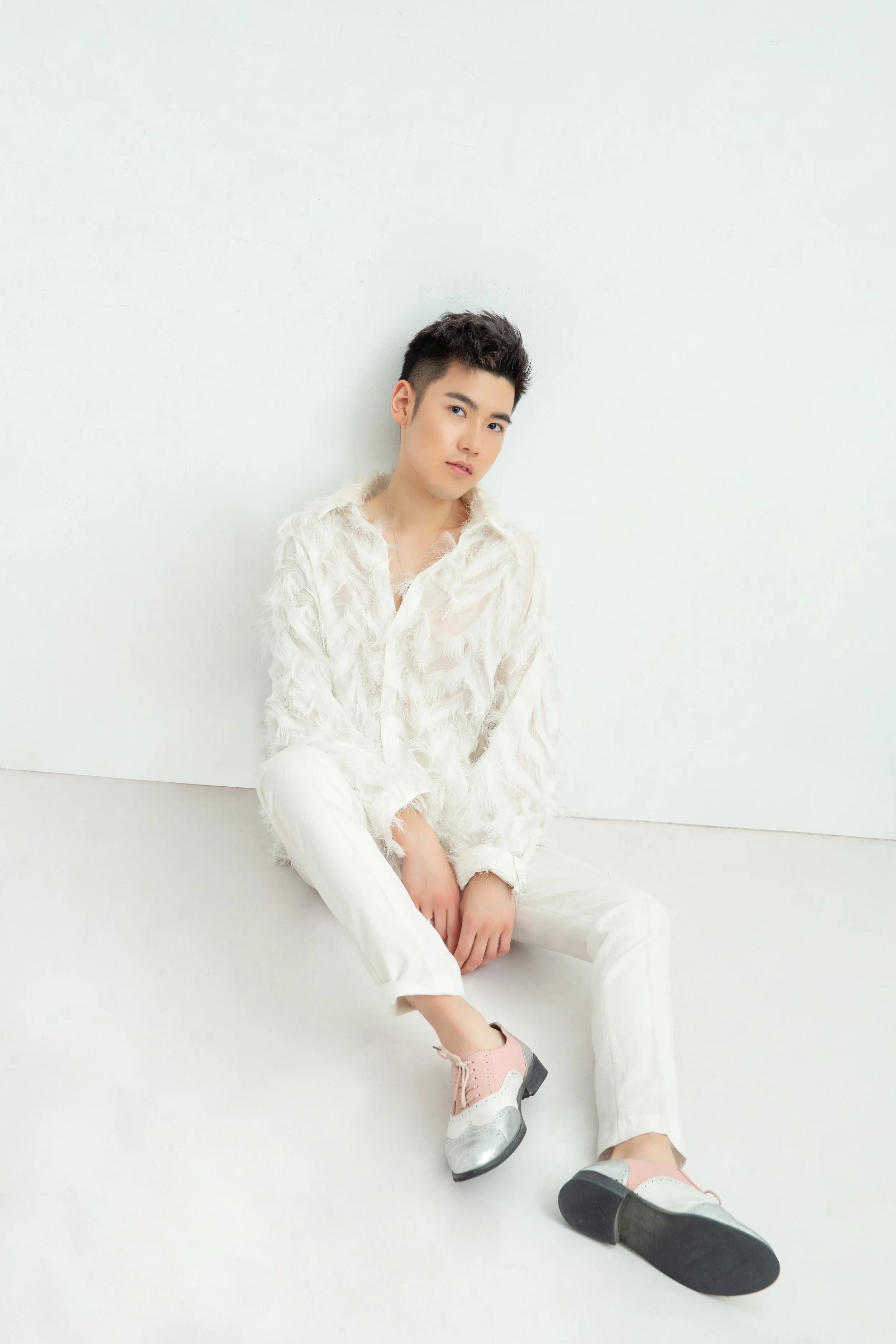 A young man with short dark hair, wearing a white feathered shirt and white pants, sitting against a plain white wall, with his left leg crossed over his right, wearing pink and white shoes, and looking at the camera.