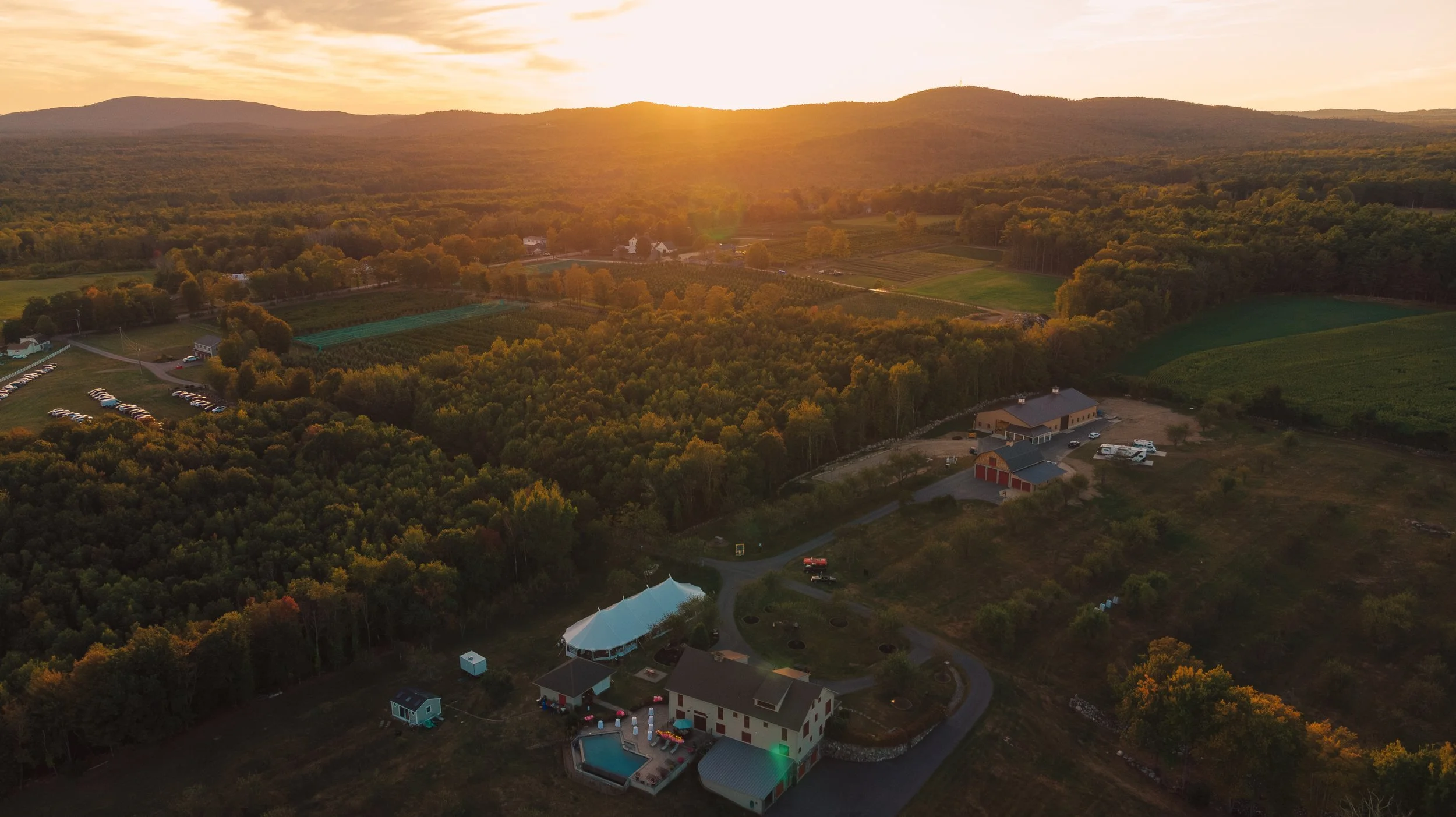A rural landscape during sunset with rolling hills, dense trees, a few buildings, a parking lot filled with cars, a swimming pool, and a tent.