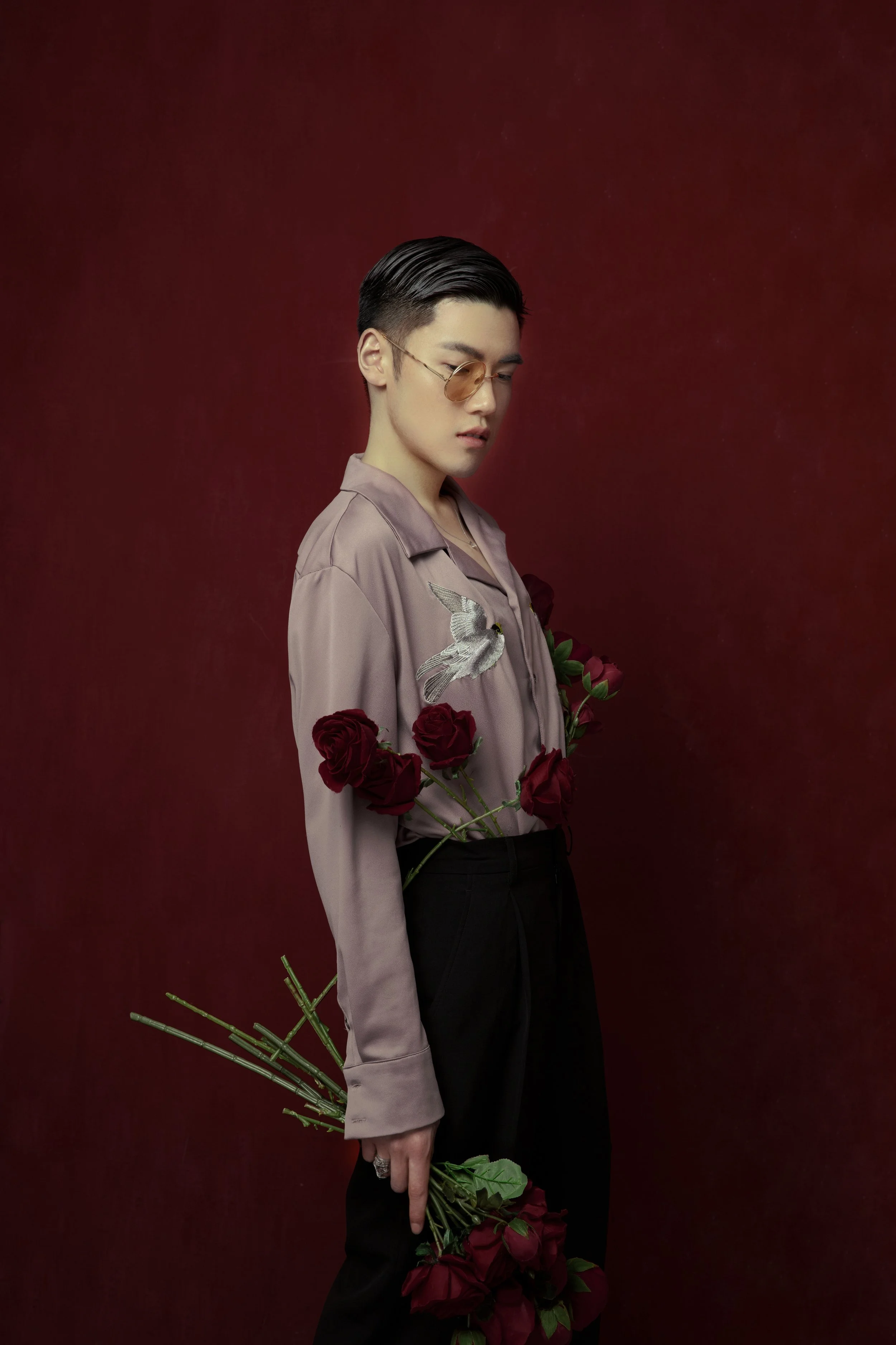 A person with short dark hair, wearing tinted glasses, a beige shirt with a bird embroidery, and dark pants, standing against a dark red background, holding red roses with some pressed against their hip and pointing downwards.