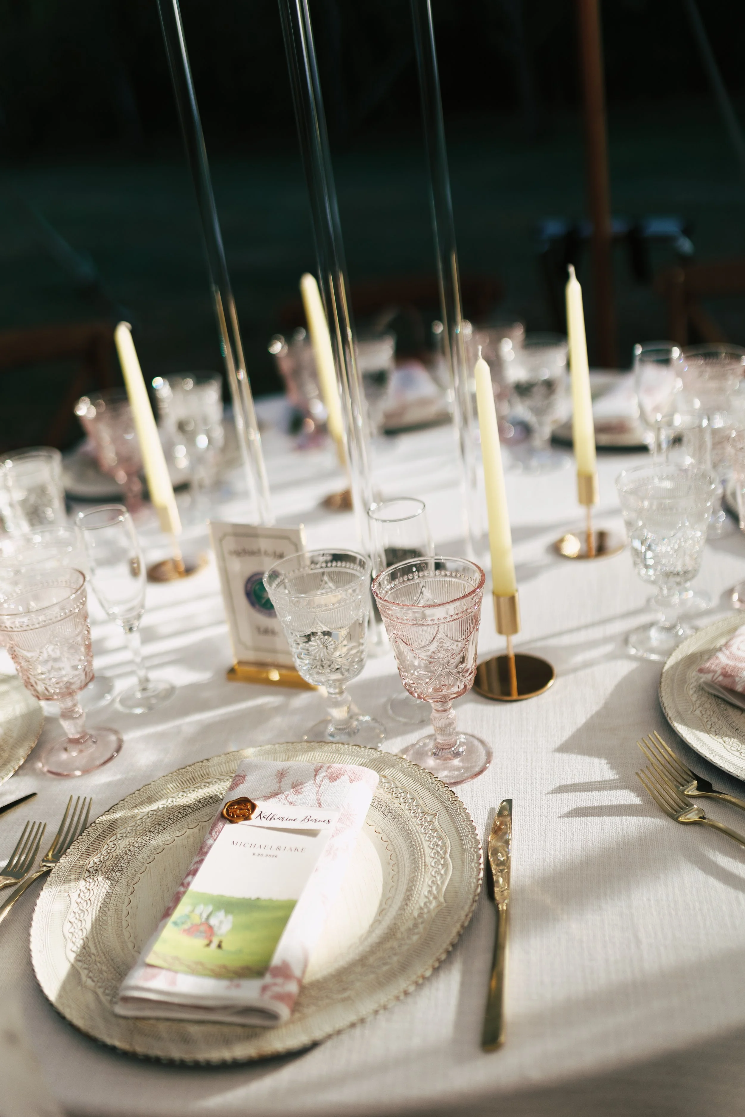 Elegant white table setting with gold accents, pink glassware, white candles, gold cutlery, and a menu with a floral design.