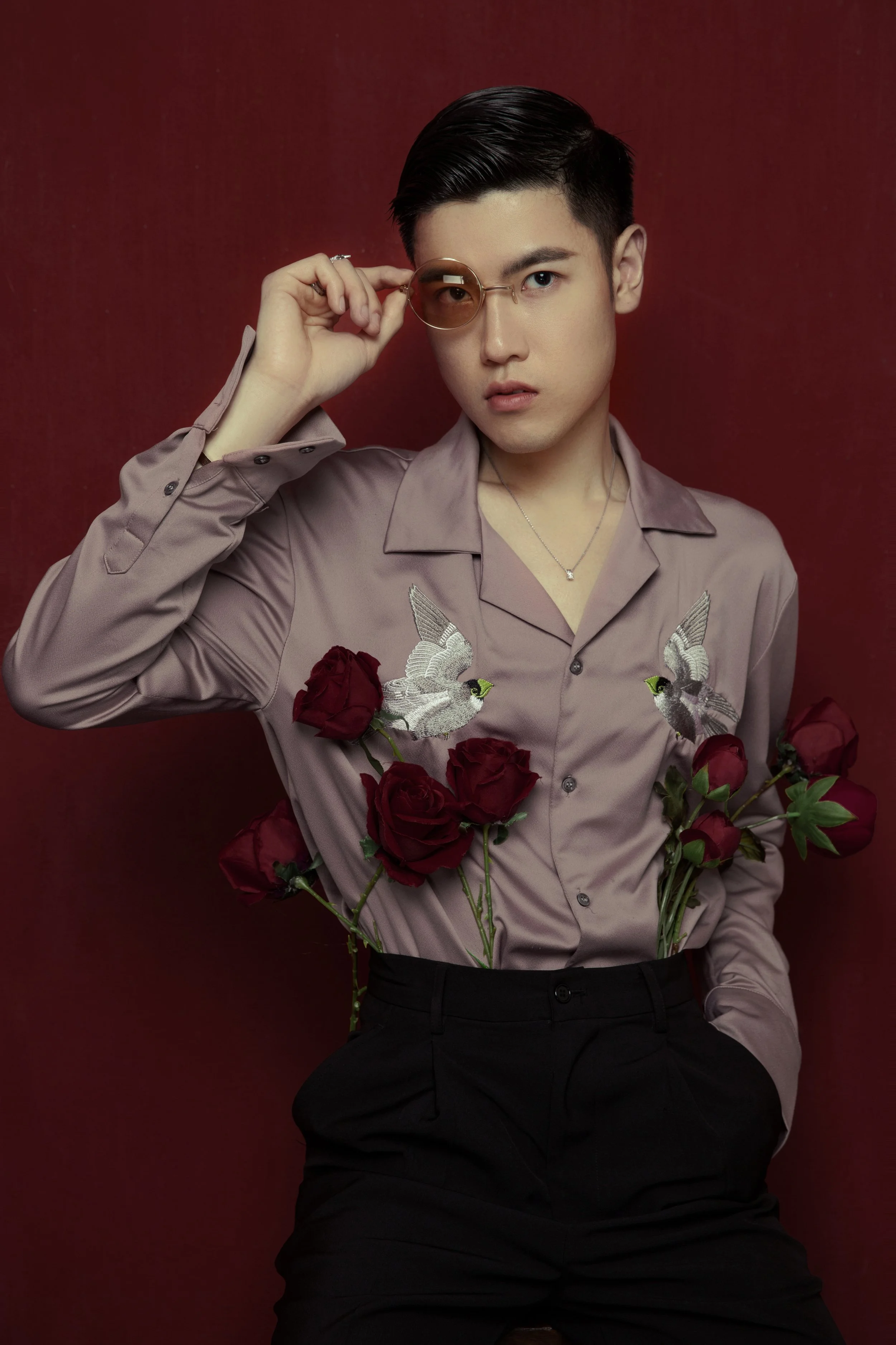 A young man with dark hair and fair skin wearing a light purple shirt with embroidery and roses, holding a pair of sunglasses, against a burgundy background.