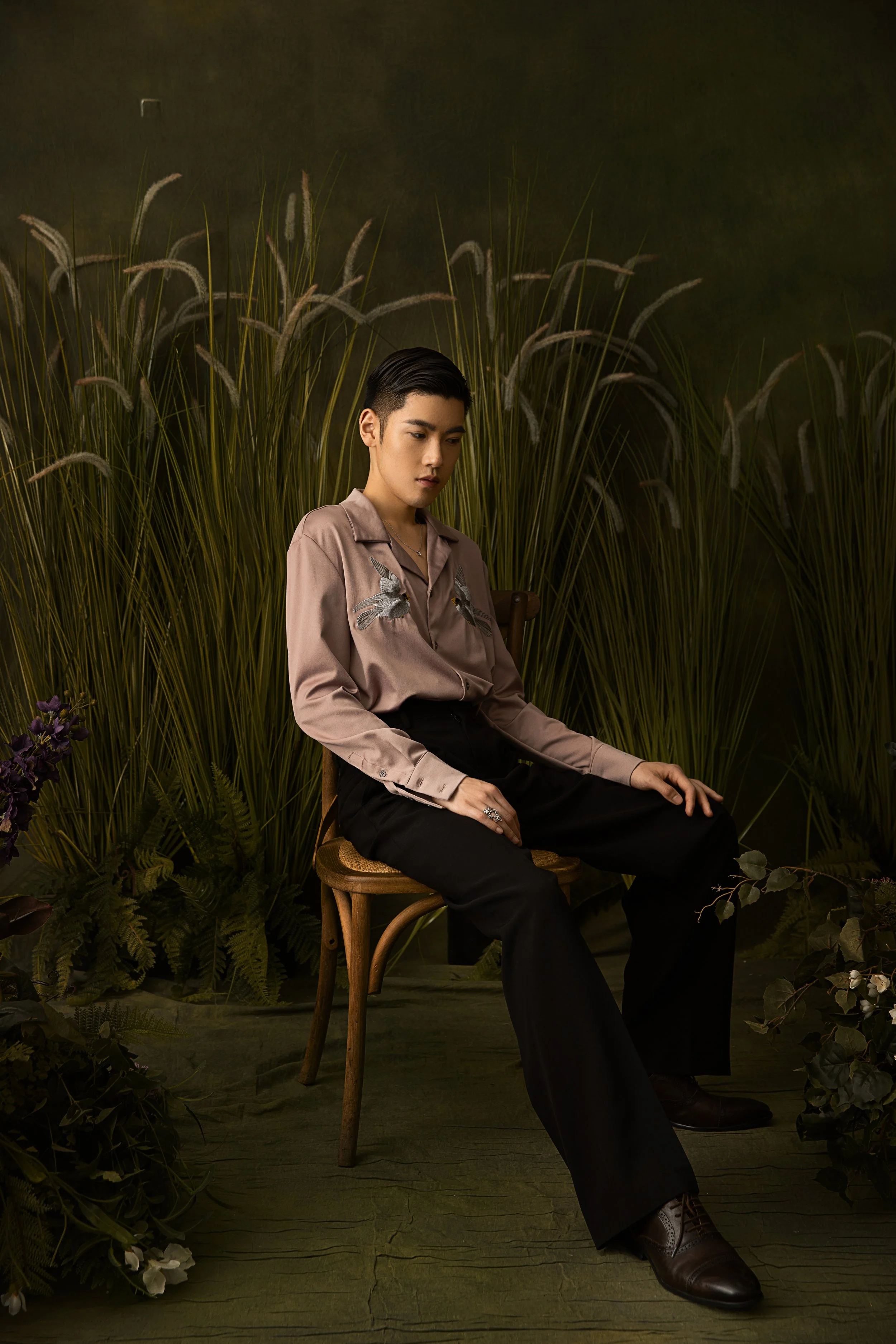 A young man with short black hair sitting on a wooden chair surrounded by green plants and dark background, wearing a beige shirt with embroidered birds and black trousers.