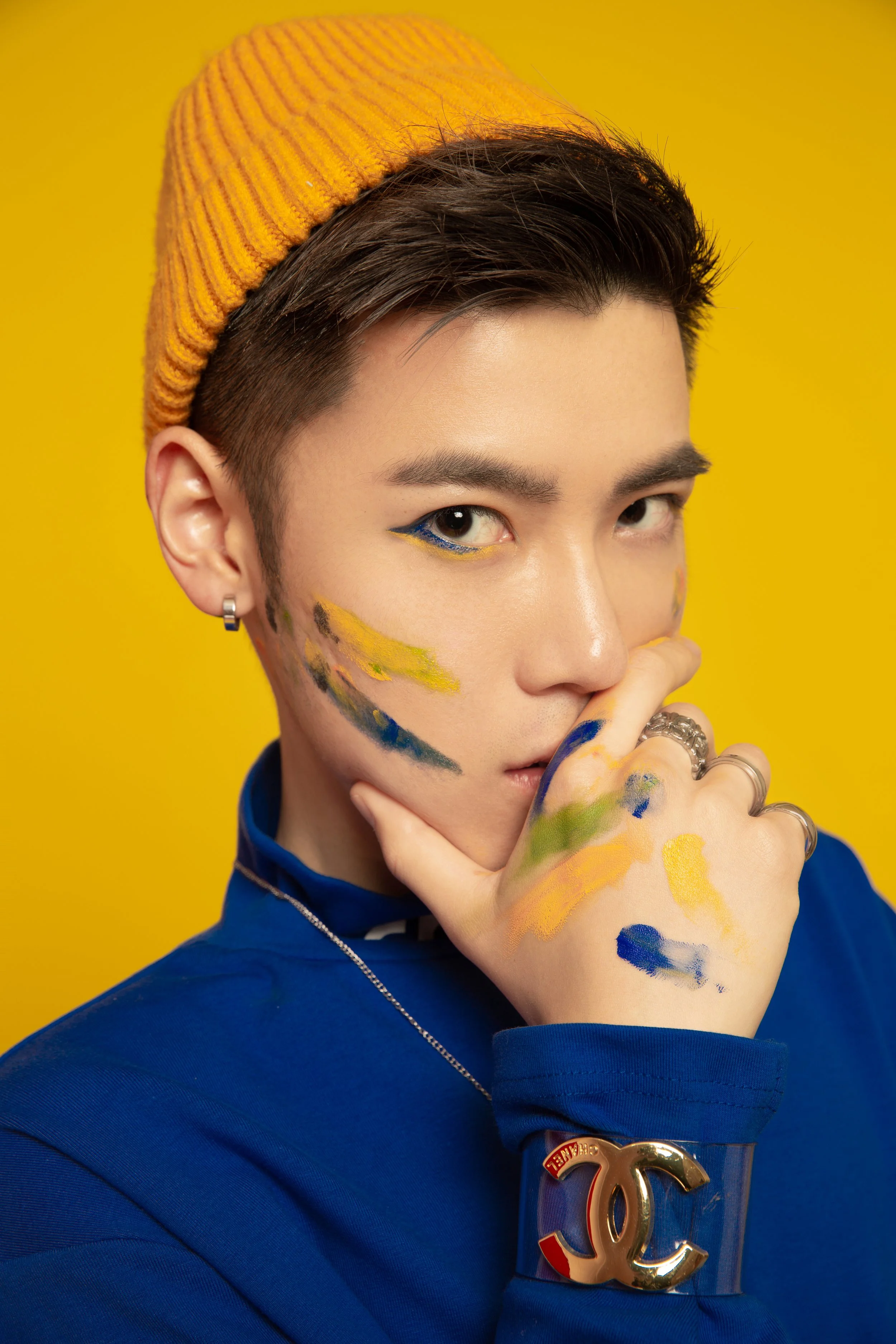 A young person with dark hair wearing a yellow knit cap, blue eye makeup, and a blue jacket with a gold Chanel logo, against a yellow background. They have paint on their face and hand, and are wearing earrings and rings.