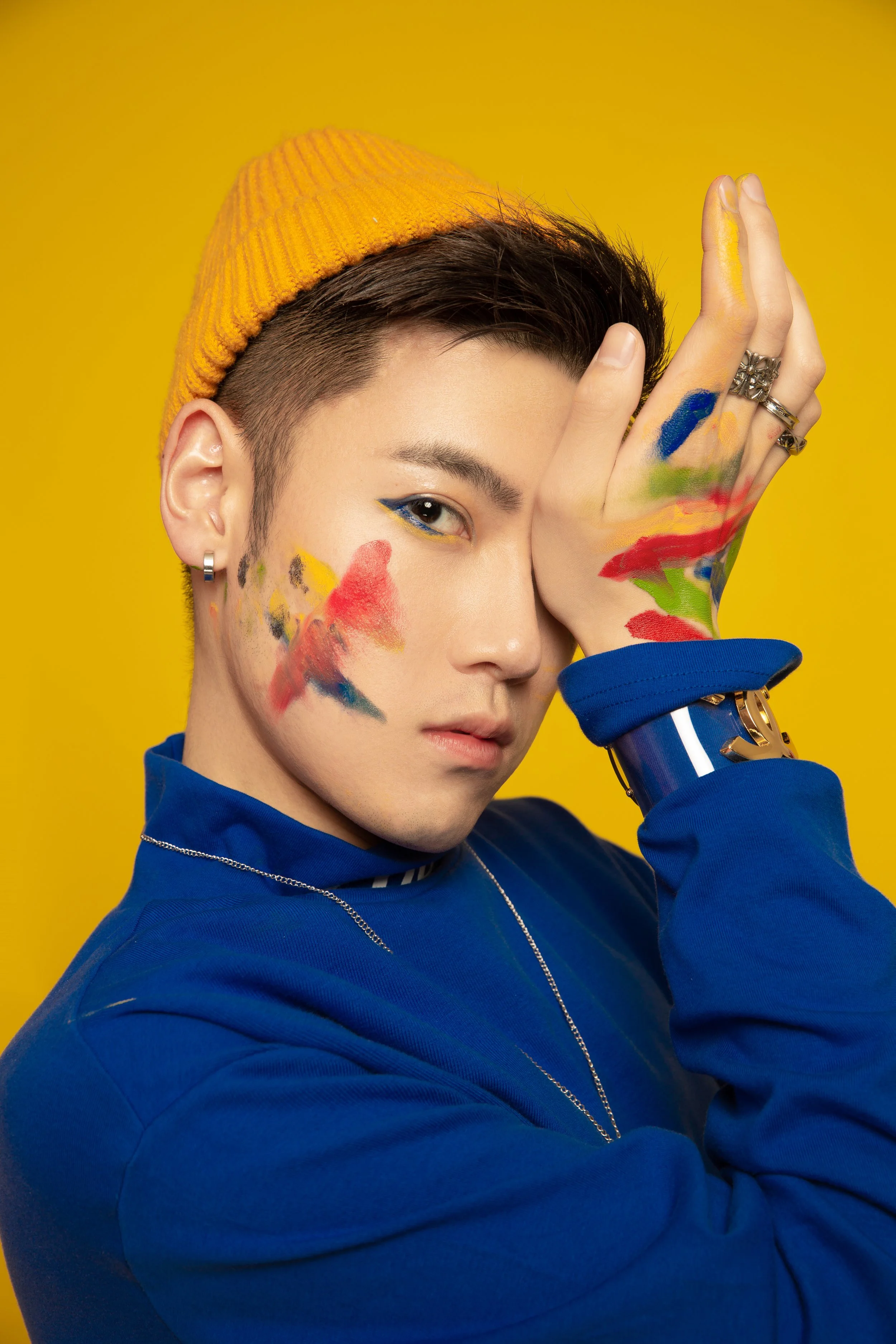 A person with short dark hair, wearing a yellow beanie and a blue jacket, poses with one eye visible and one hand covering part of their face, against a bright yellow background. They have colorful paint on their face and hand.