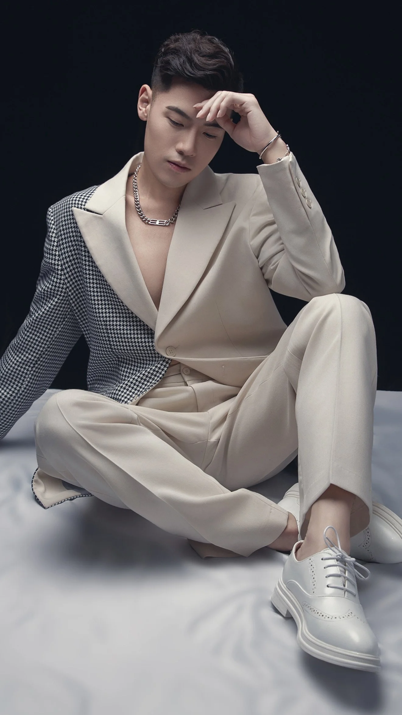A young man sitting on a white surface against a black background, wearing a beige suit with one checkered sleeve, white sneakers, and silver jewelry, with his hand on his forehead.