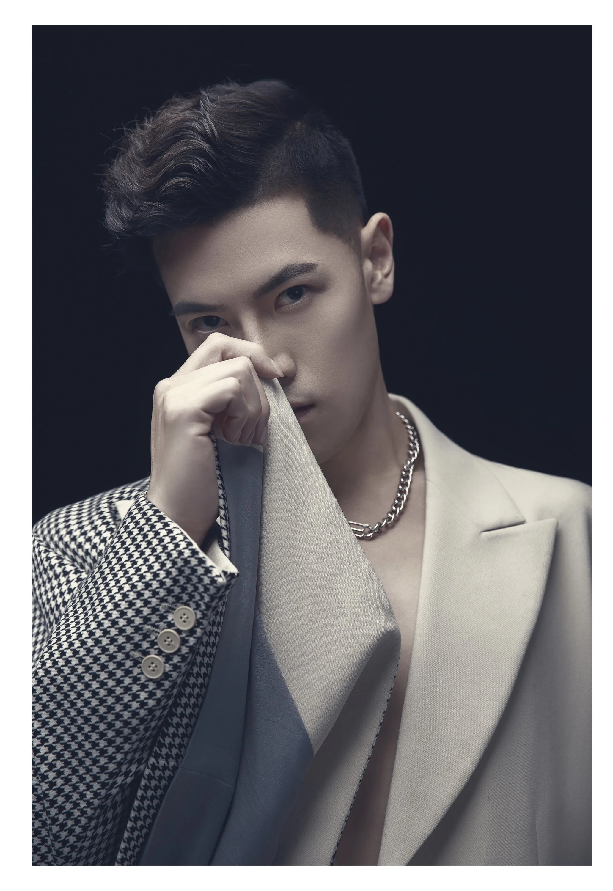 A young man with styled dark hair, wearing a beige jacket over a patterned shirt, accessorized with a silver chain necklace, holding part of his jacket near his face, against a dark background.