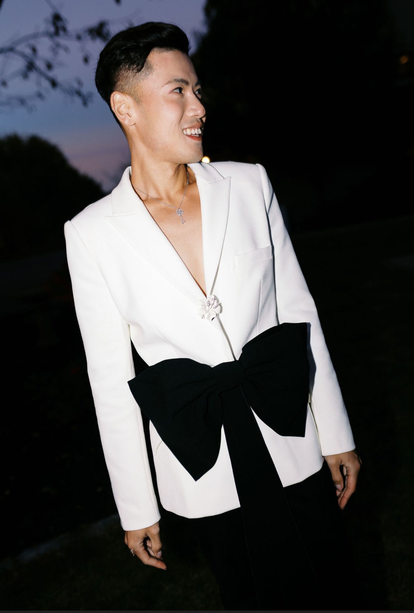 A woman in a white blazer with a large black bow on the front, smiling and looking to the side, outdoors at dusk or night.