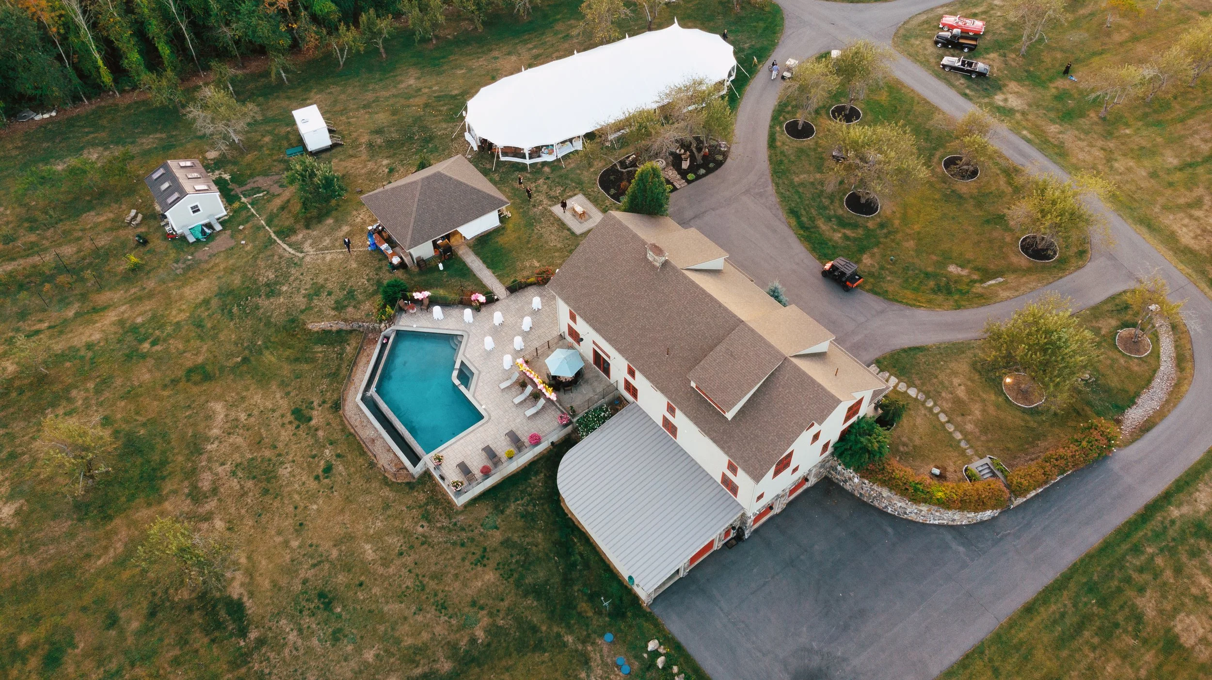 An aerial view of a backyard with a swimming pool, a large white tent, several parked cars, a house, and a landscaped yard with trees and pathways.