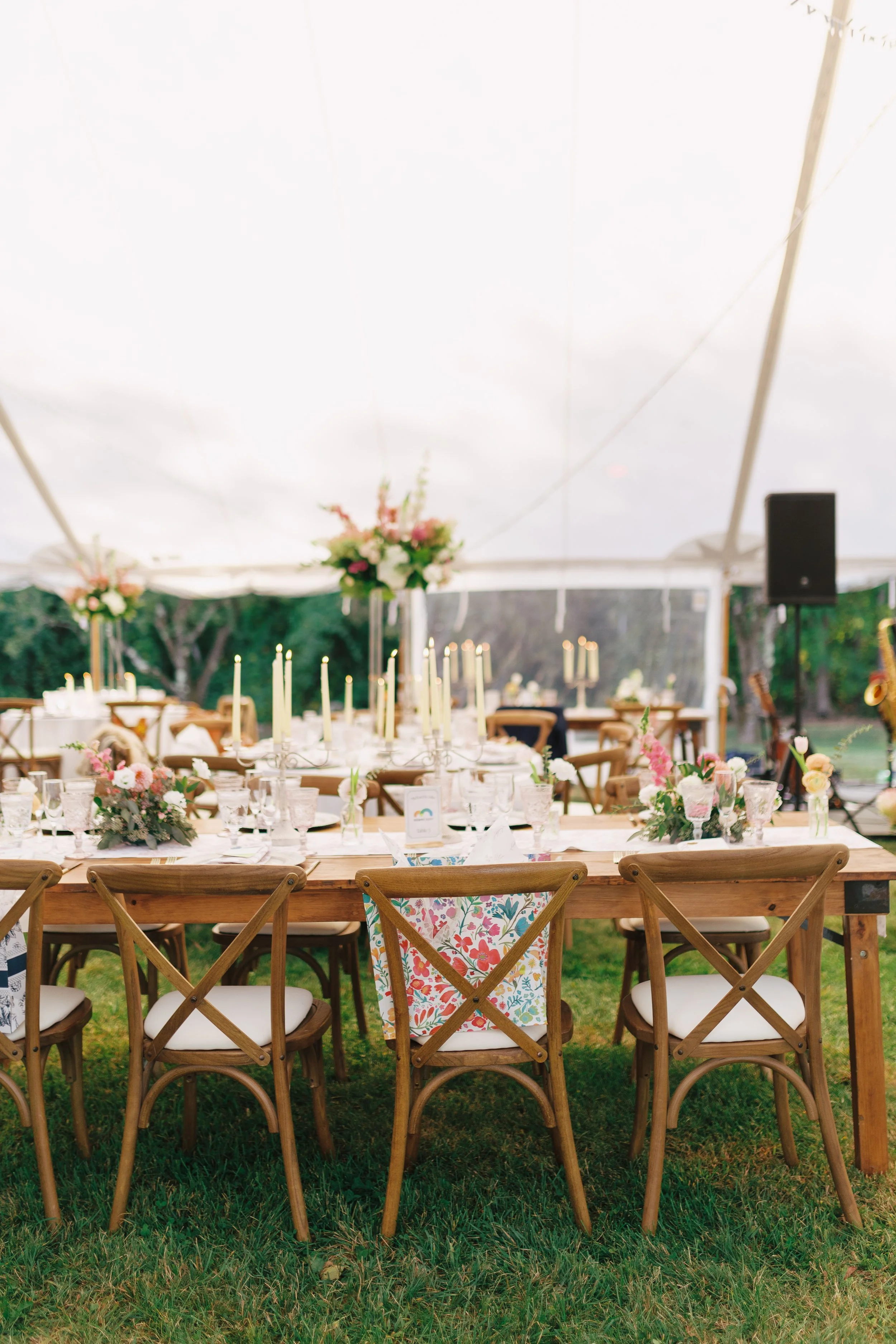 Wedding reception under a white event tent with decorated tables, floral centerpieces, candles, and chairs with patterned seat cushions, set on green grass.