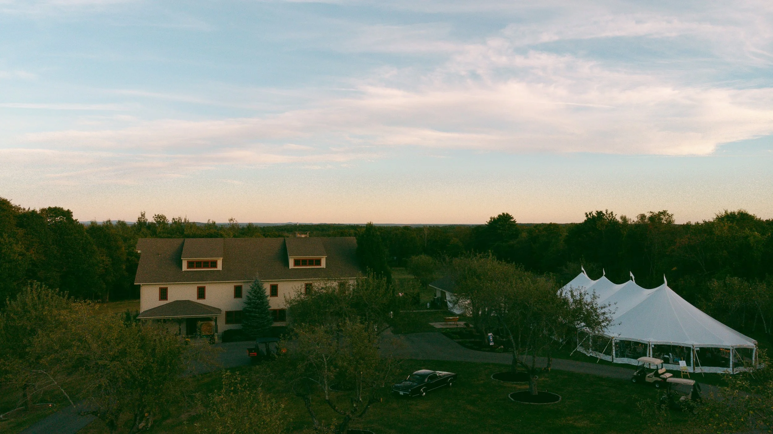 A suburban area with a house, trees, and a large white event tent, under a partly cloudy sky at sunset.