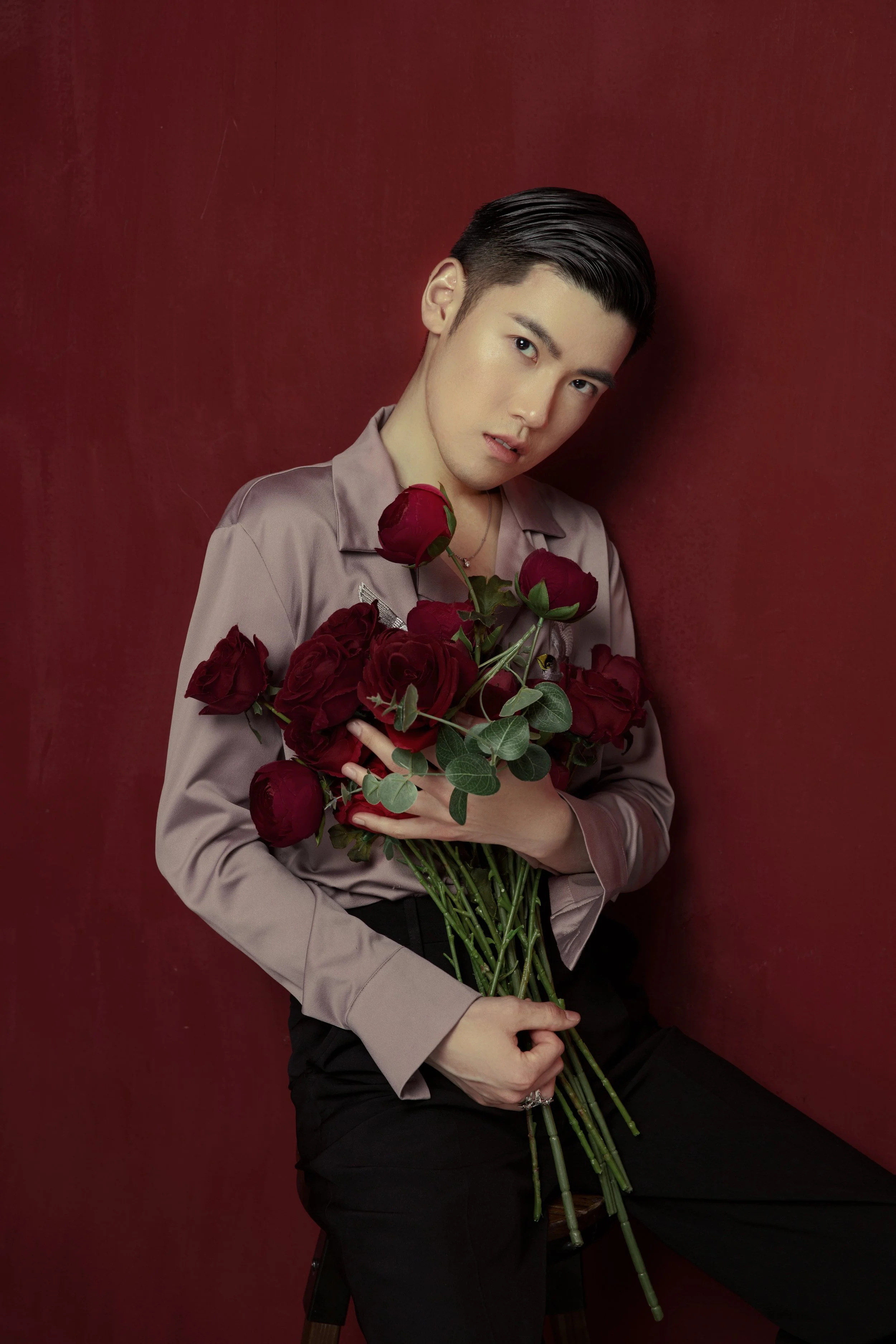 A young man with short, slicked-back hair holding a bouquet of red roses against a deep red background.